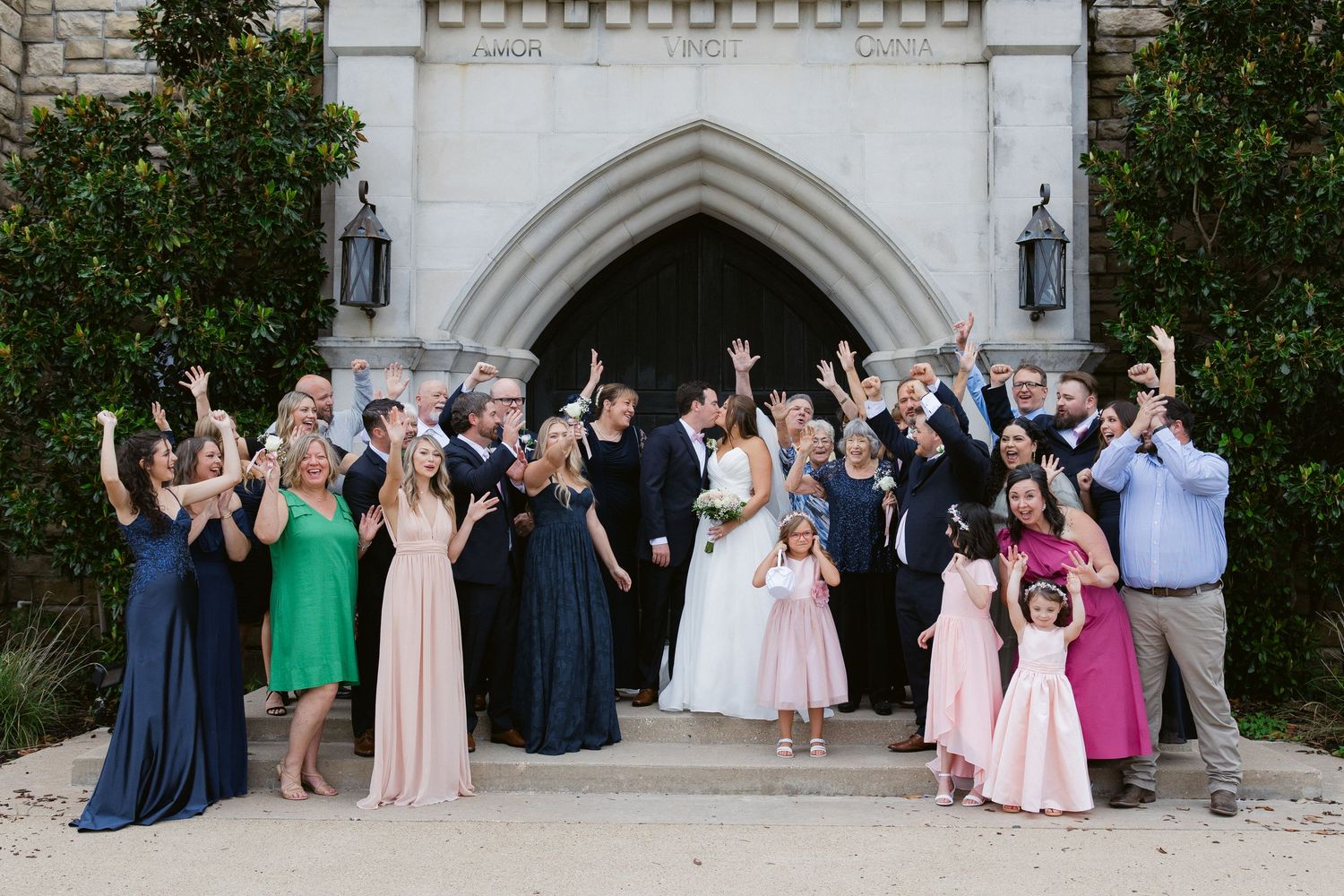 A large group of wedding guests pose together for a formal photo outside a white stone church with arched doorway.