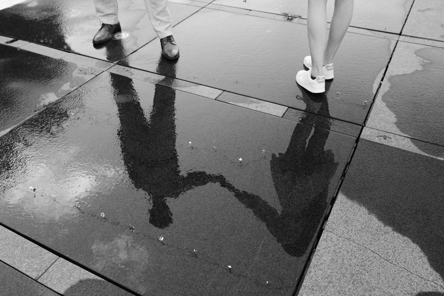 Black and white photo of two people walking on wet pavement with reflections visible in rain puddles.