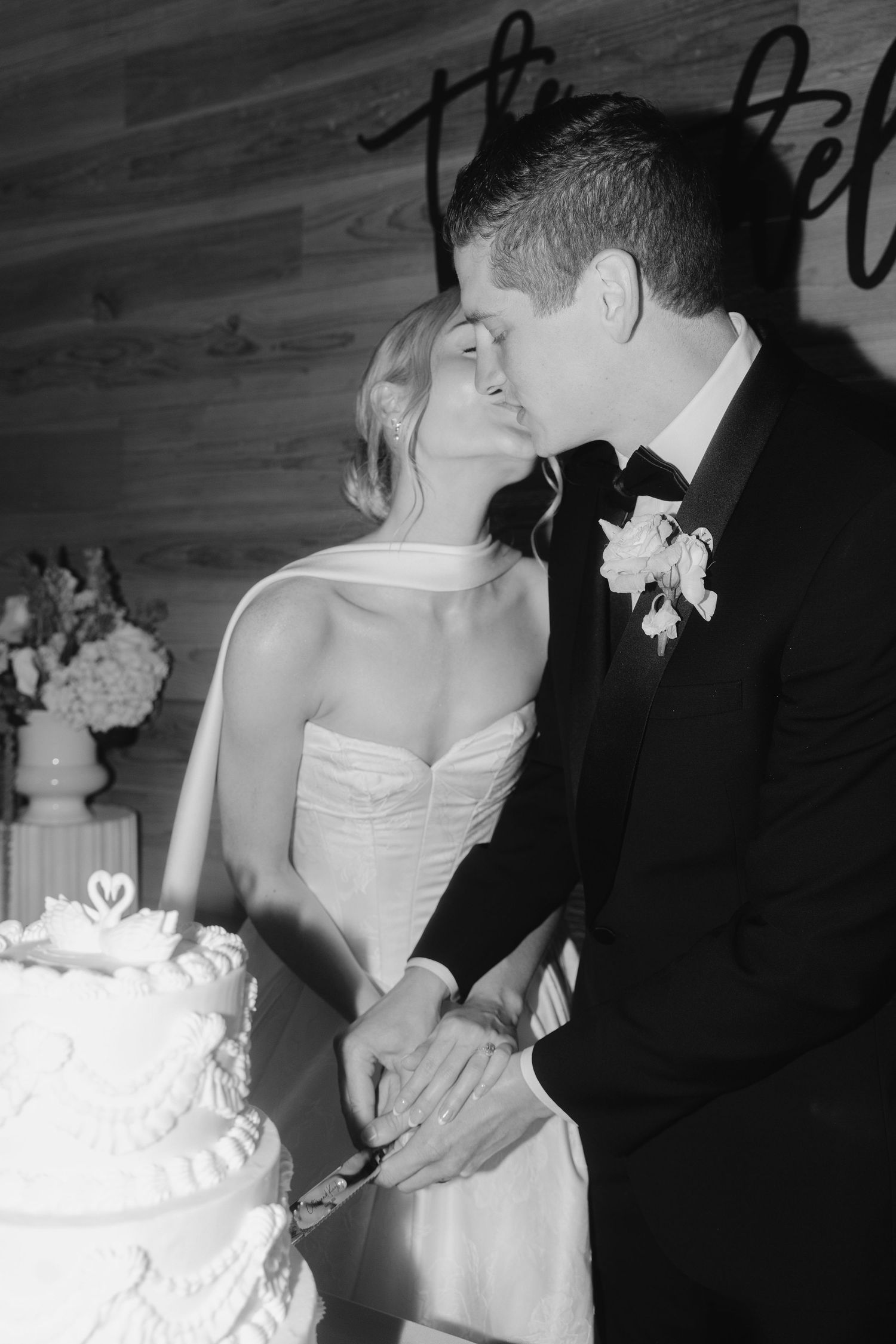 Black and white photo of bride and groom kissing while cutting wedding cake at reception.