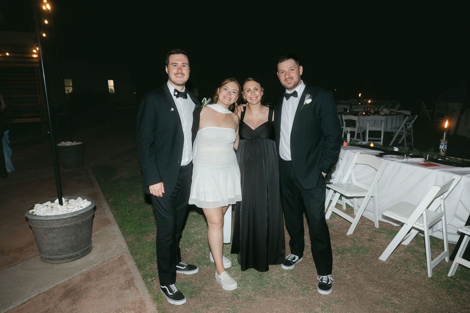 Two couples in formal attire pose together outdoors at a wedding reception at night.
