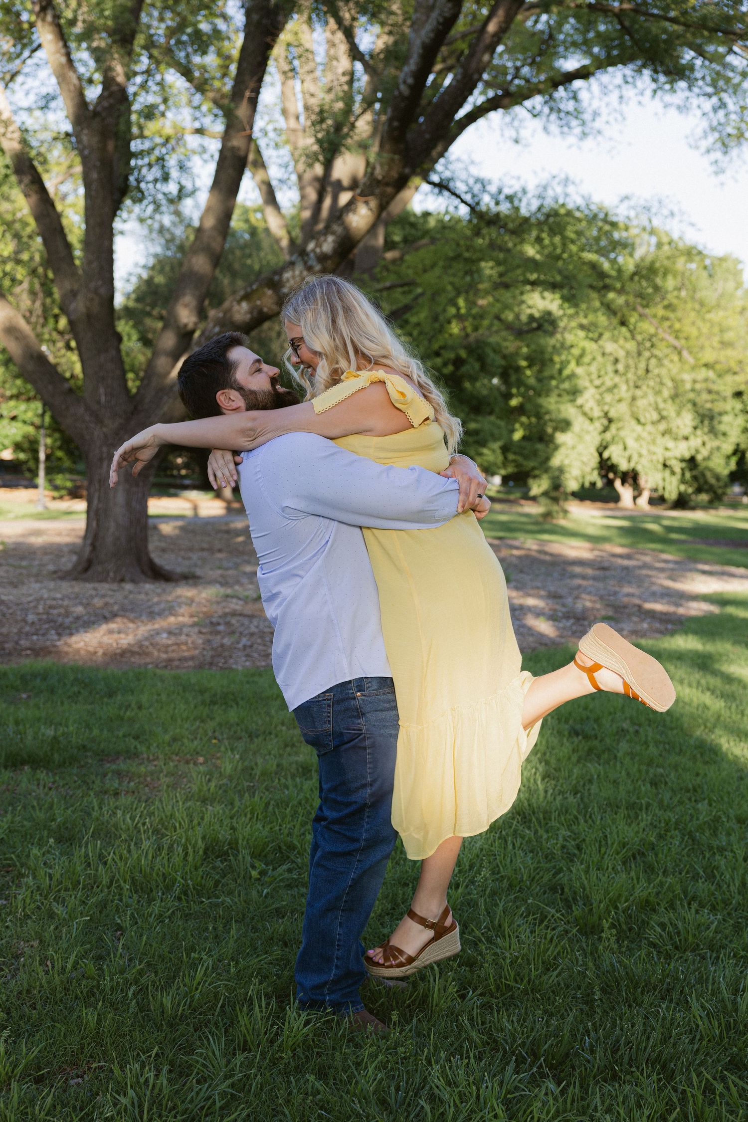 A romantic embrace in a park setting with a yellow sundress and casual attire against a backdrop of green trees.