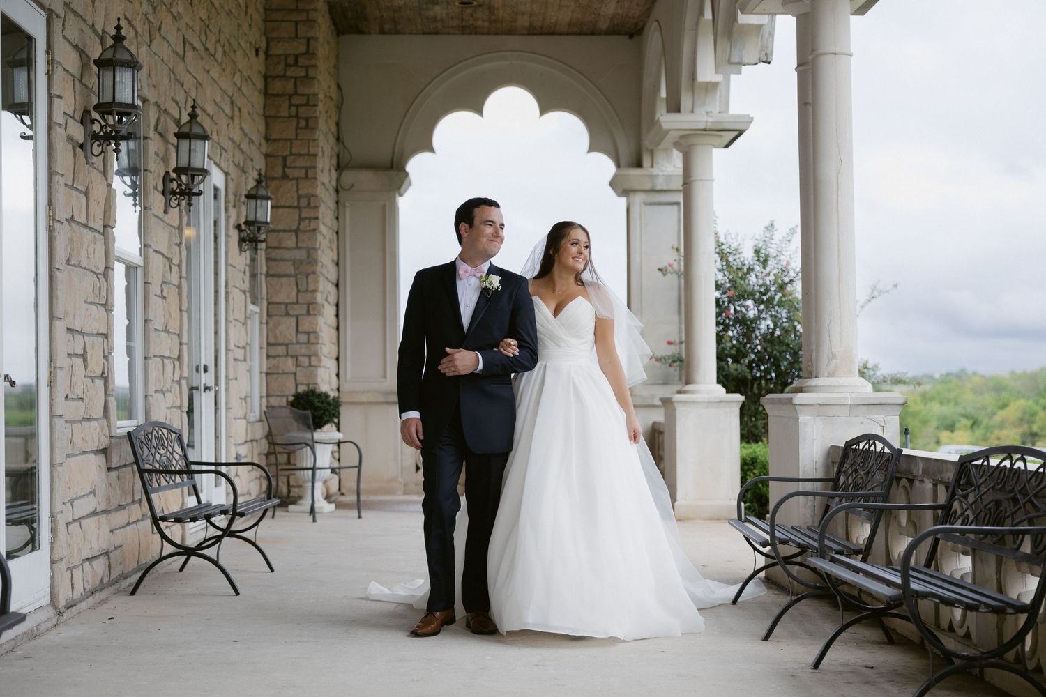 Elegant wedding couple stands under stone archway of venue overlooking scenic countryside view.