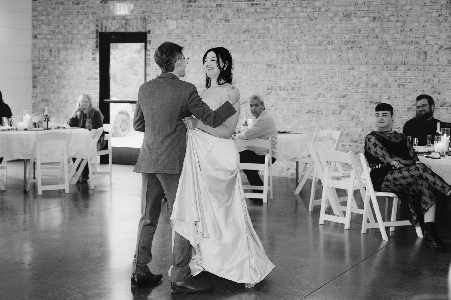 A couple shares their first dance in a rustic reception venue with exposed brick walls and white chairs.