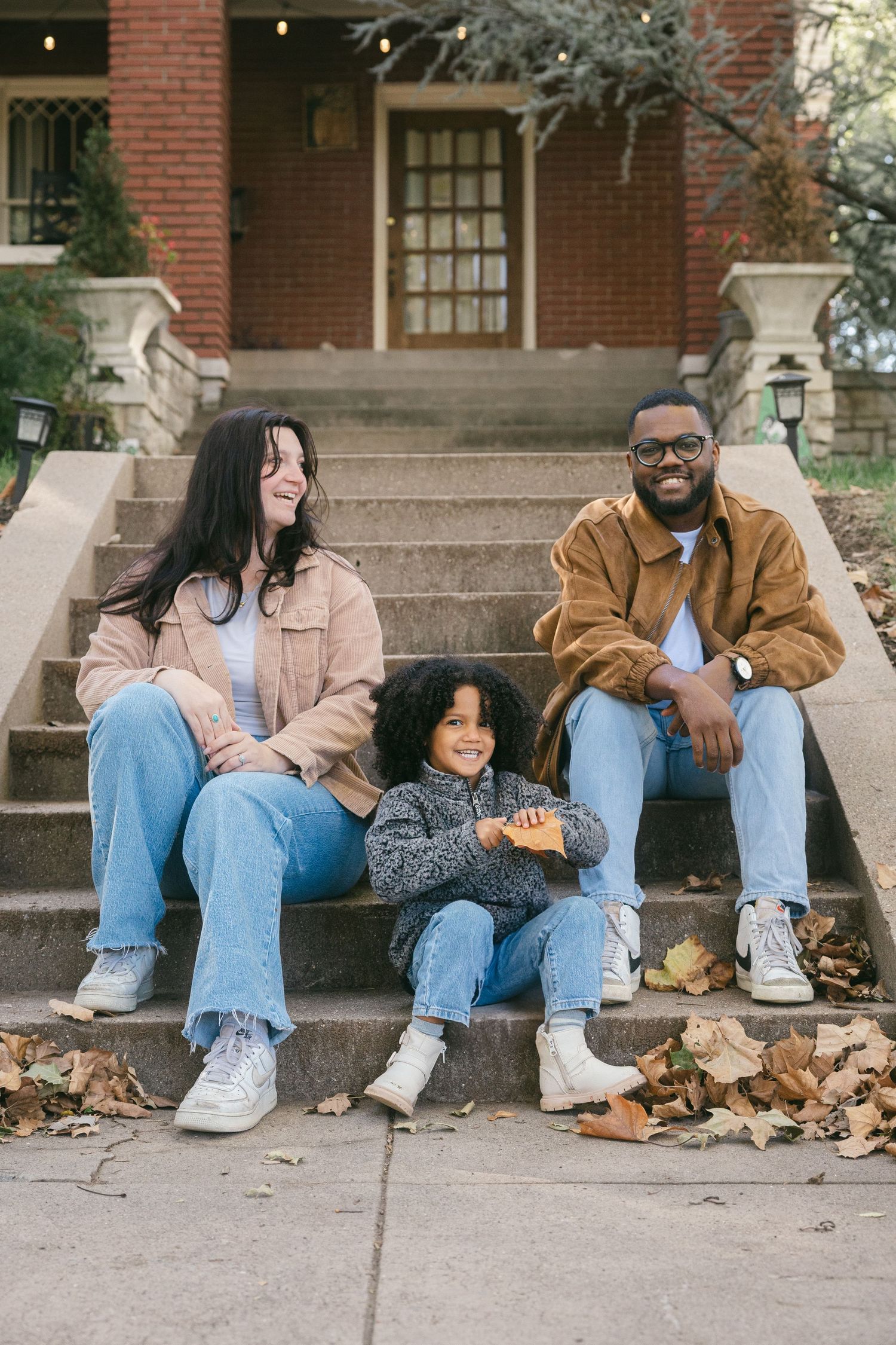 A couple sits casually on brick steps surrounded by fallen autumn leaves on a sunny day.