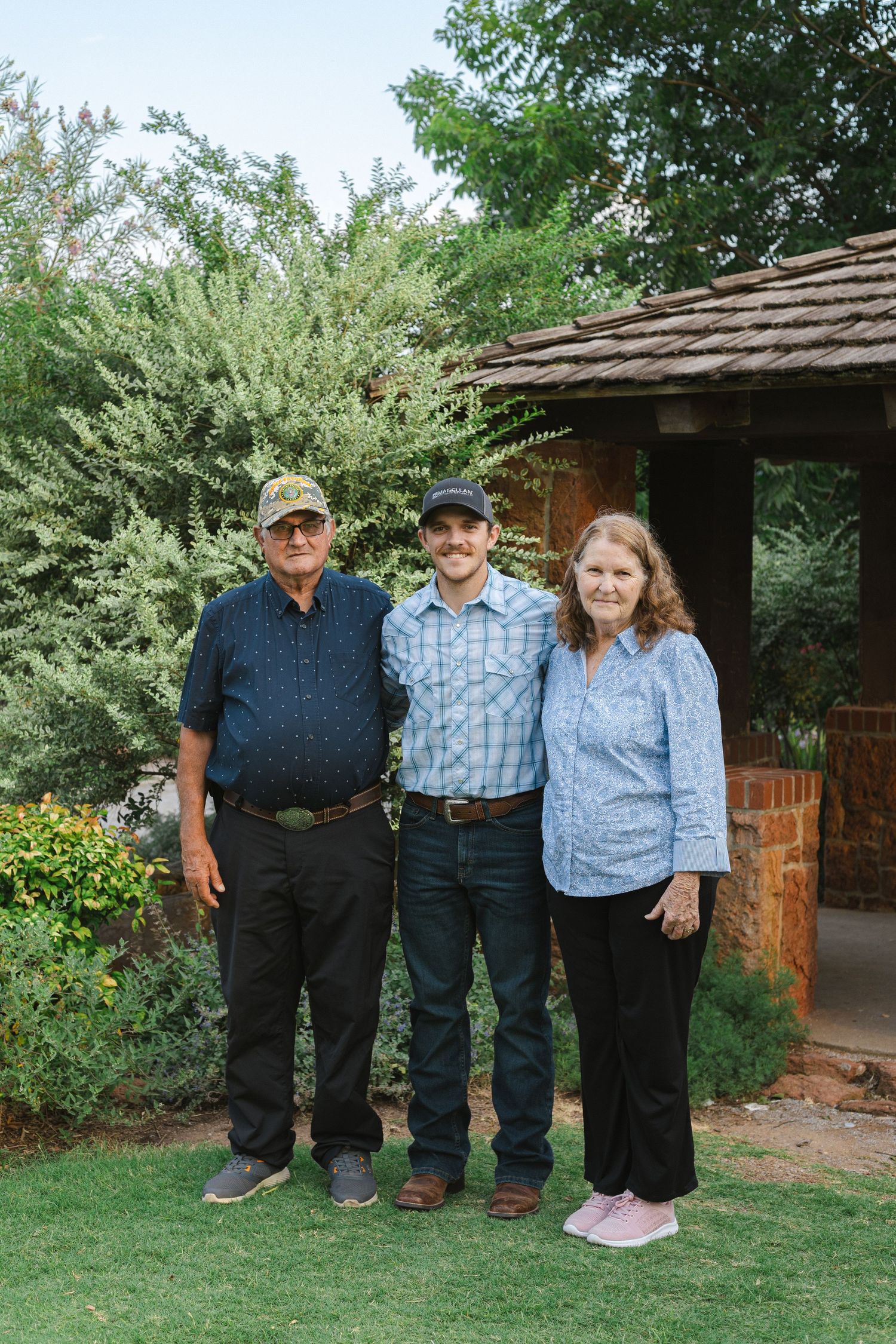 Three people stand together in casual attire outside a rustic cabin with garden landscaping.