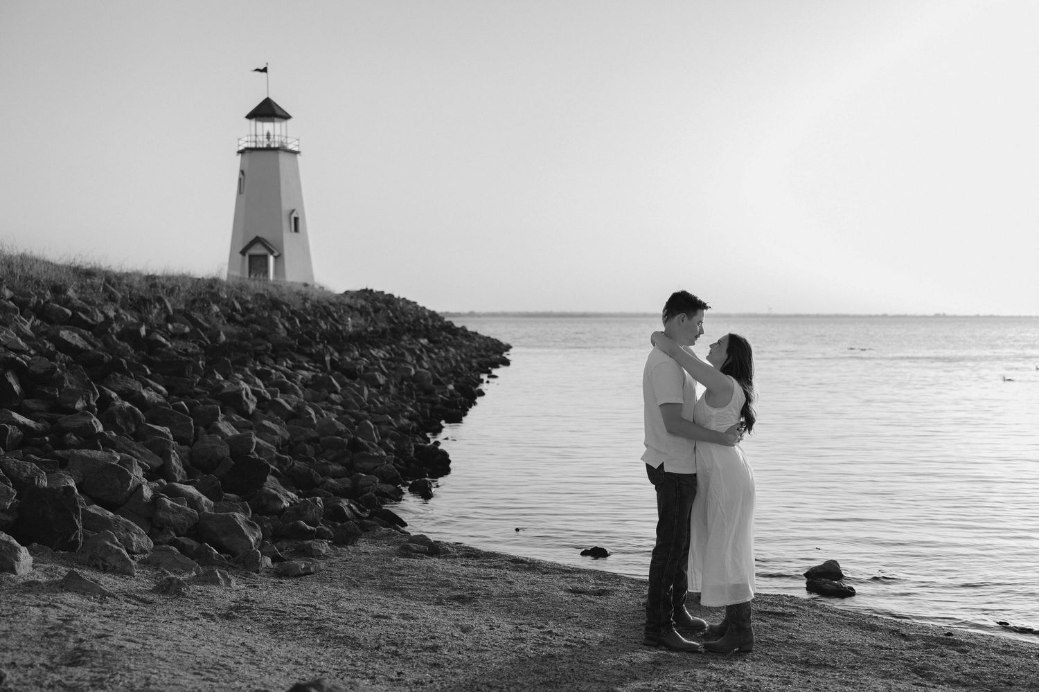 Black and white photo of a couple embracing near a lighthouse on a rocky shoreline at sunset.