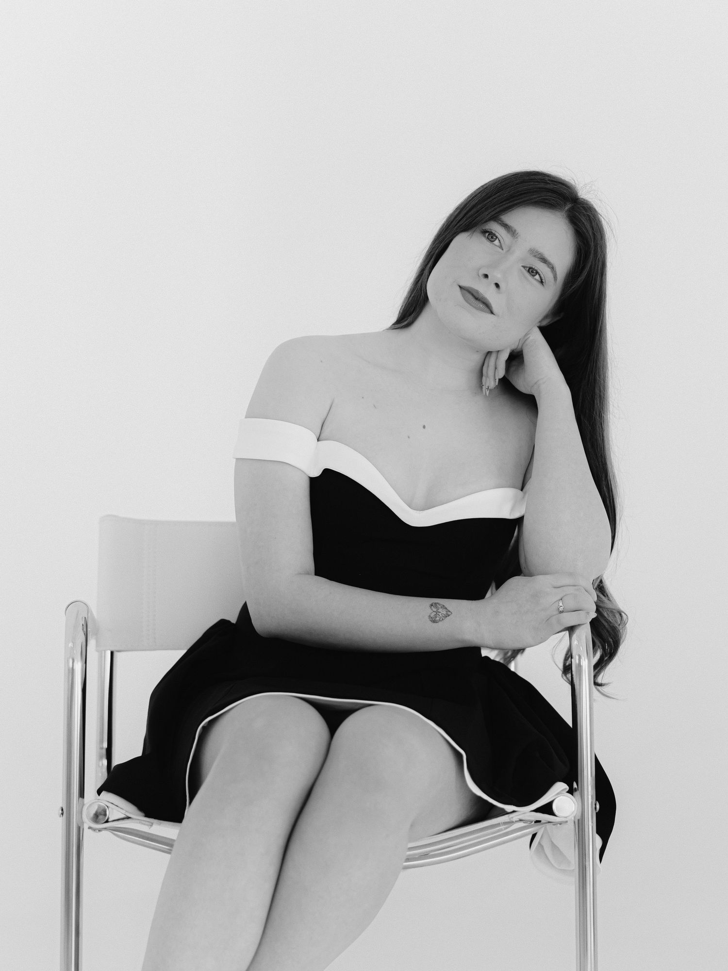 Black and white photo of a woman in an off-shoulder dress seated gracefully on a modern chair.