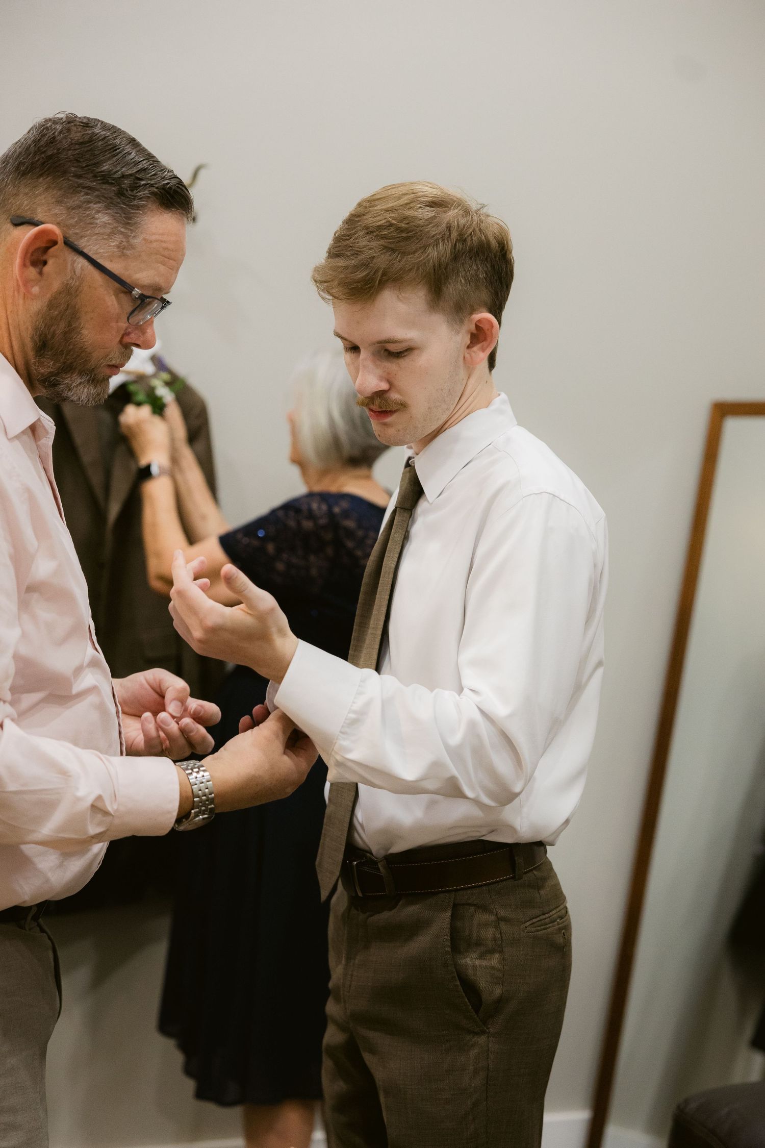 Two men getting dressed in formal attire examine their cuffs and sleeves in an indoor setting.