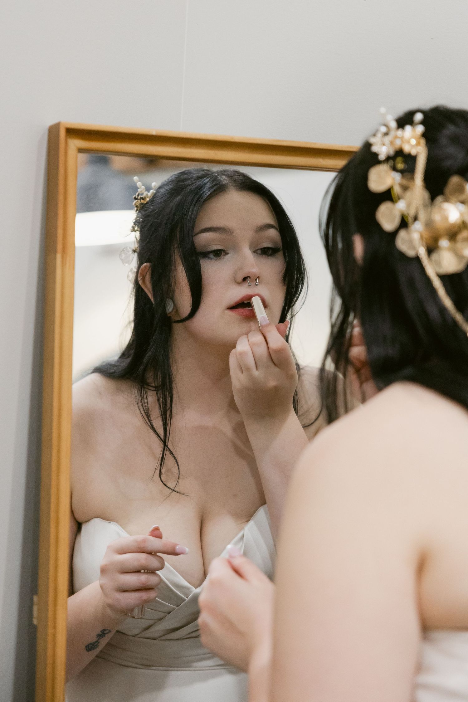 A bride applies lipstick while looking in a mirror, wearing a white dress with delicate details.