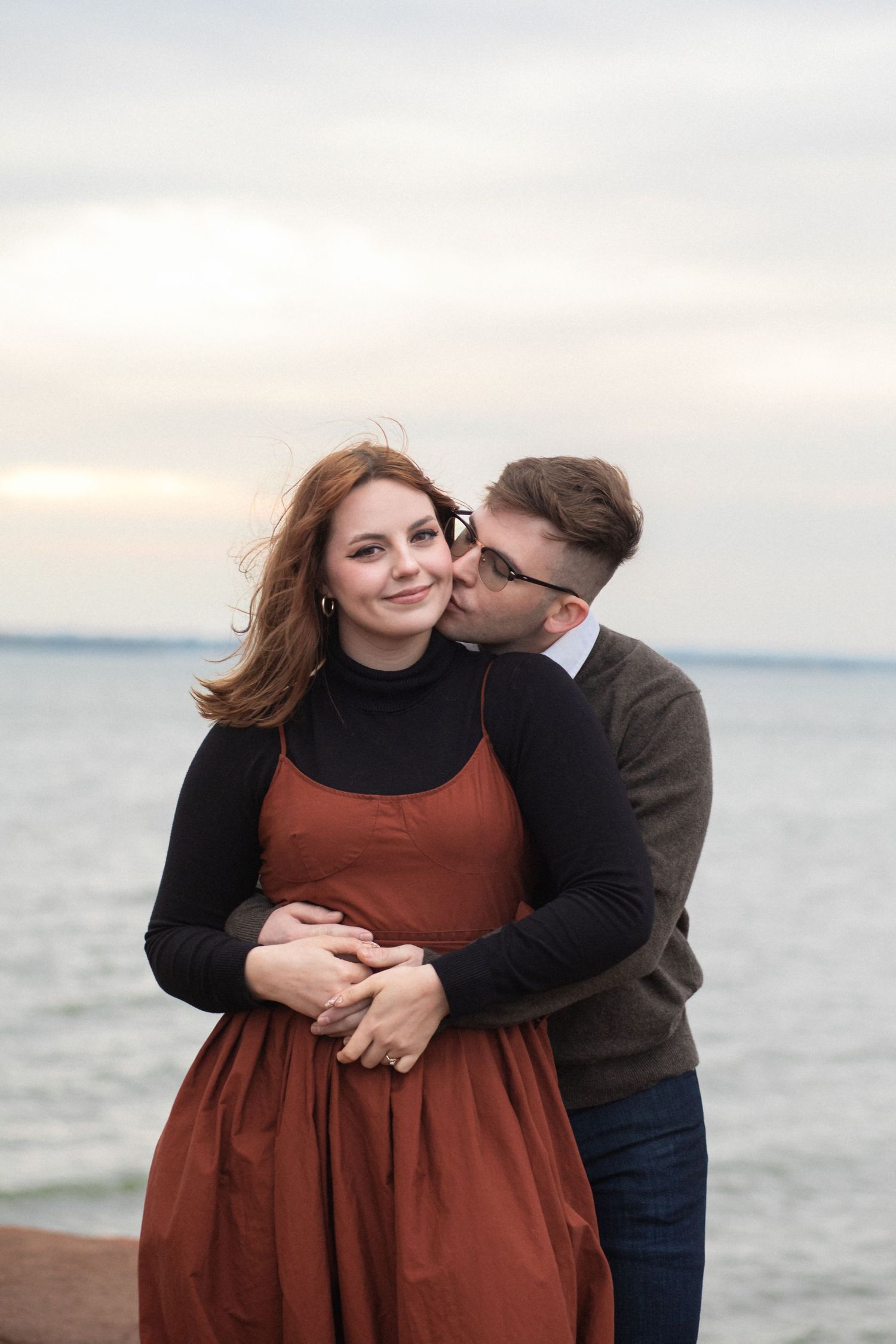 Couple embraces on a peaceful beach at sunset, with the water and cloudy sky creating a romantic atmosphere.