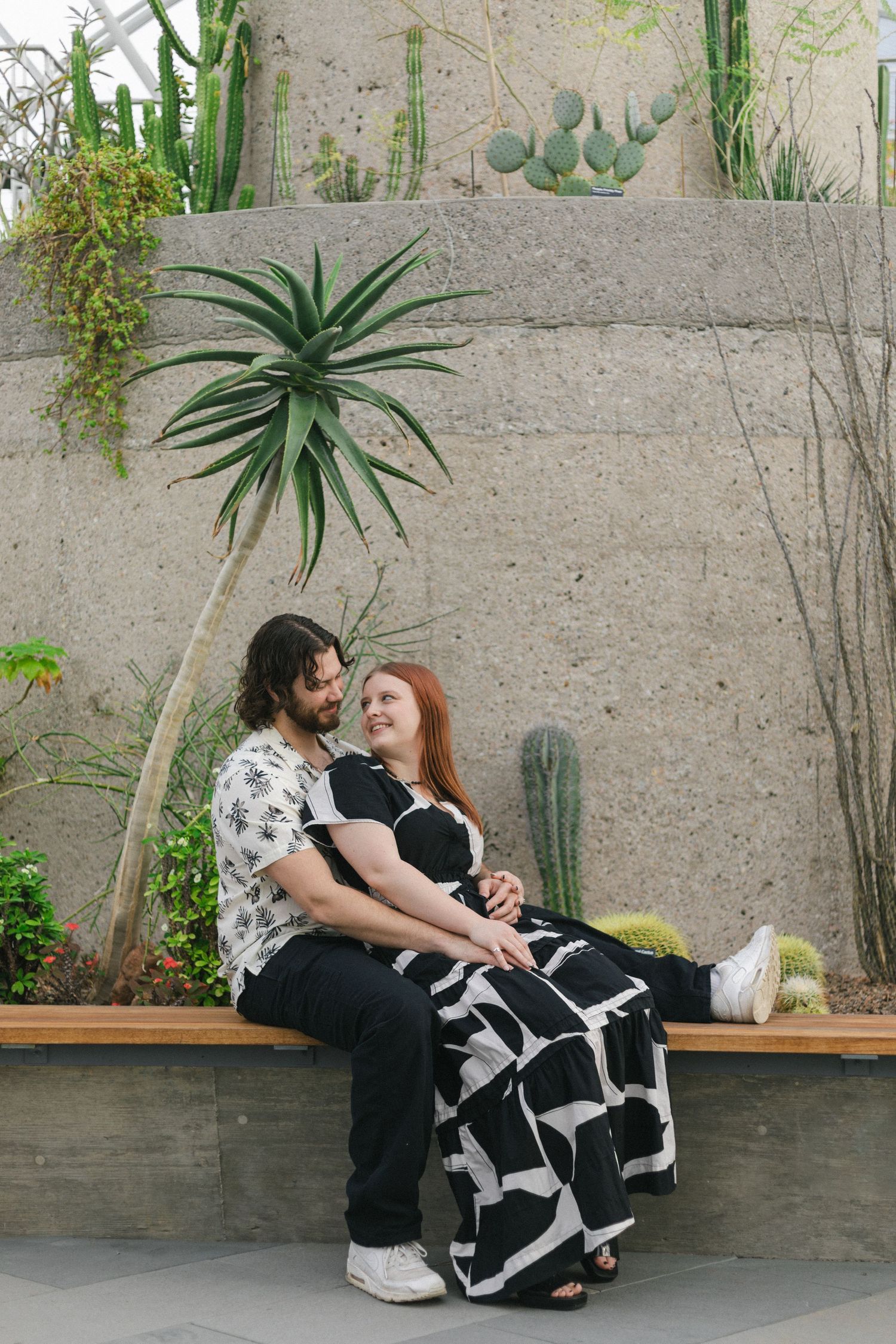 A couple shares an intimate moment on a bench against a concrete wall decorated with succulents and plants.