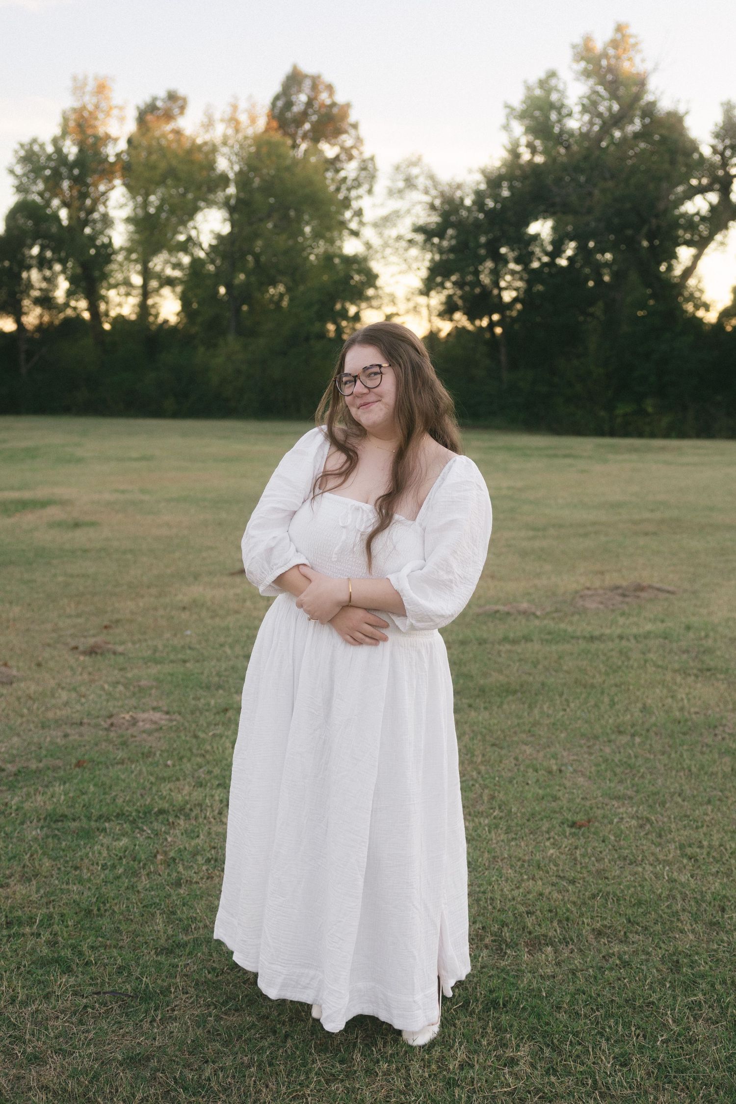 A person in a flowing white dress poses gracefully in a grassy field at sunset.