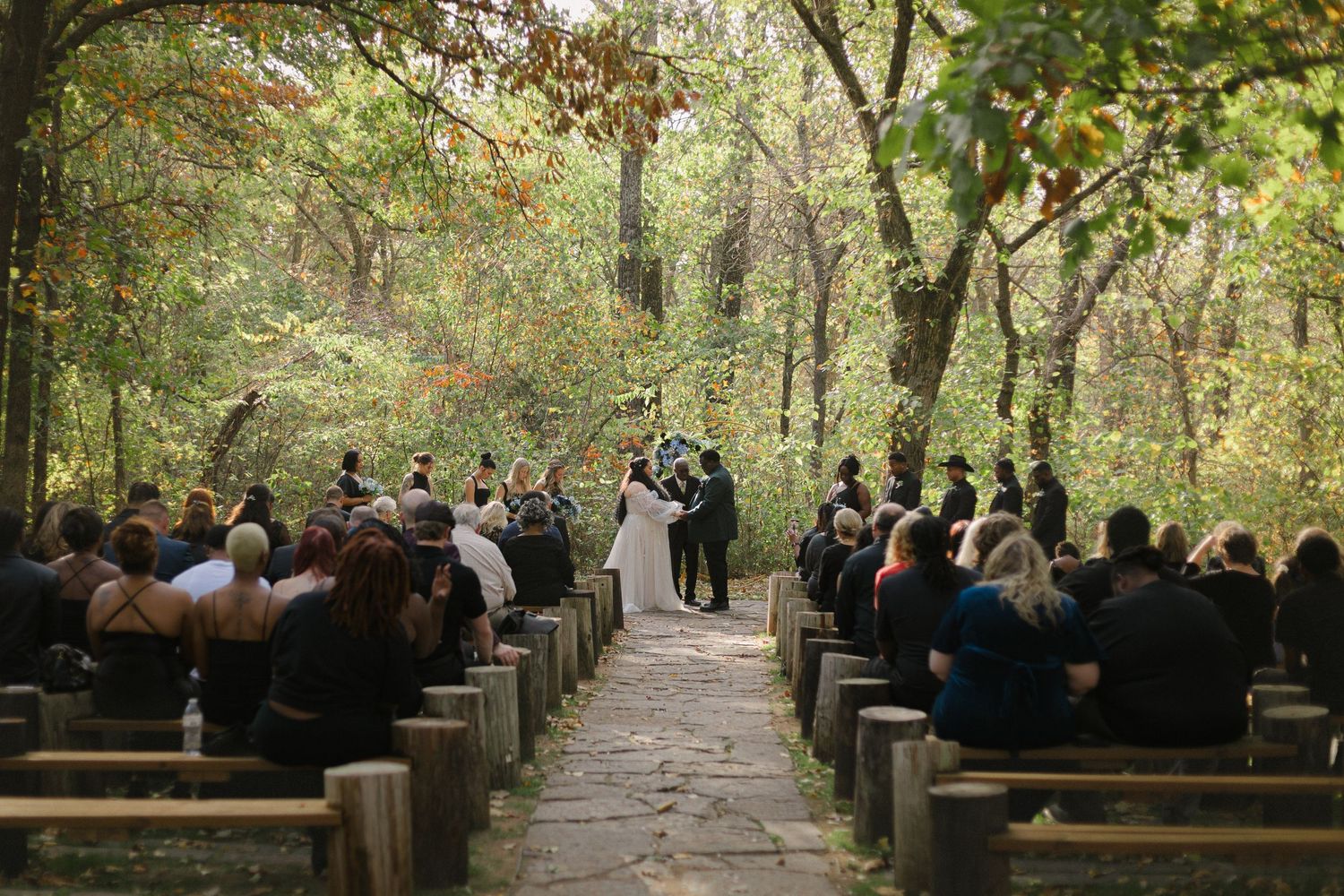 Guests seated on wooden benches along a tree-lined path during an outdoor wedding ceremony in a wooded setting.