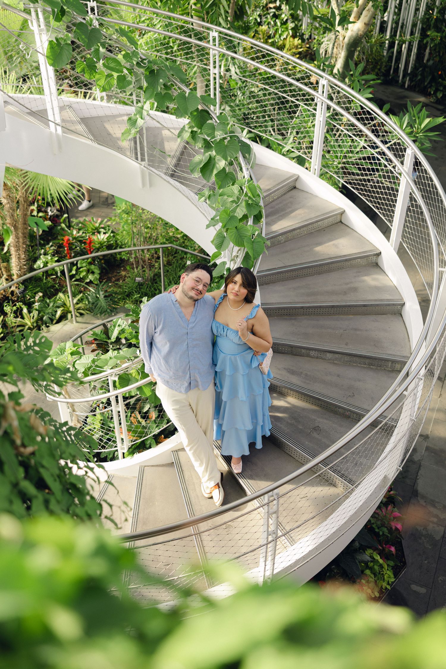 Couple posing together on a white spiral staircase surrounded by lush tropical greenery inside a botanical garden.
