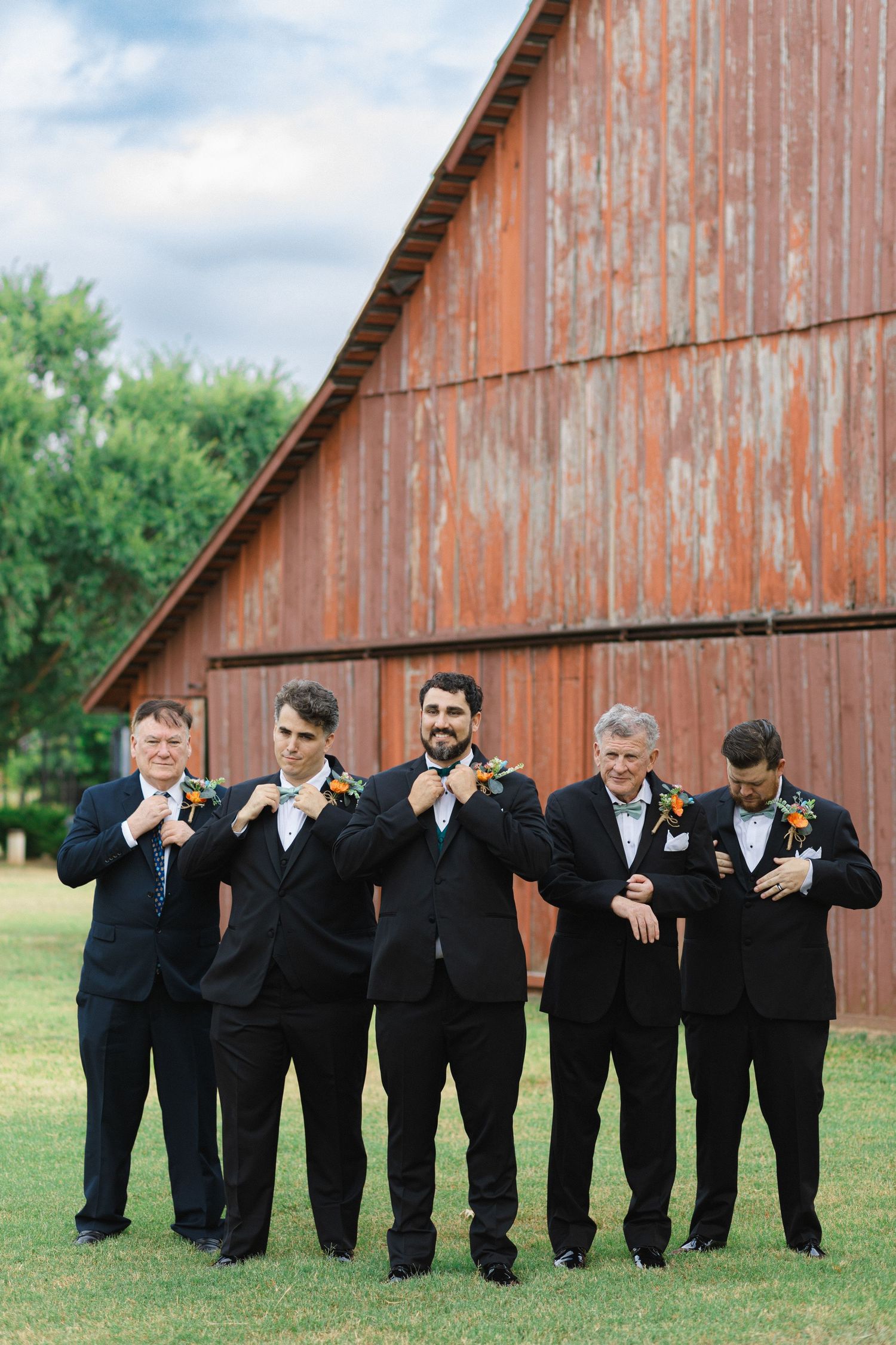 A group of groomsmen in black suits pose playfully in a line against a rustic red barn backdrop.