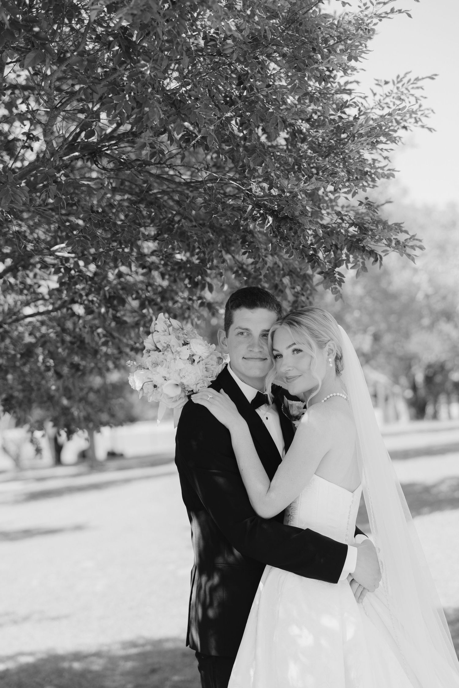 Black and white photo of bride and groom embracing under a tree on their wedding day.
