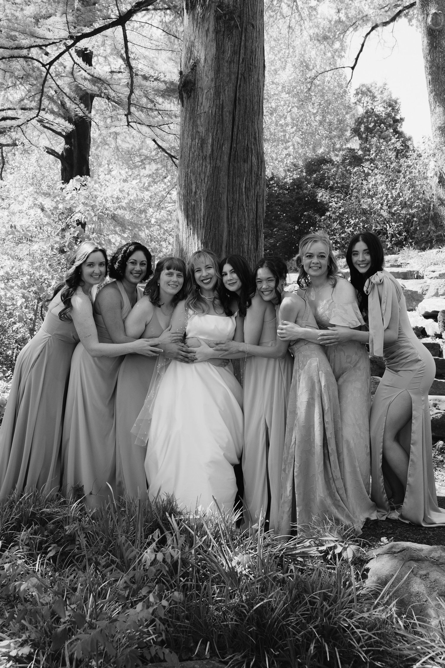 Bride in white gown surrounded by bridesmaids in mismatched dresses hugging near a large tree in black and white.