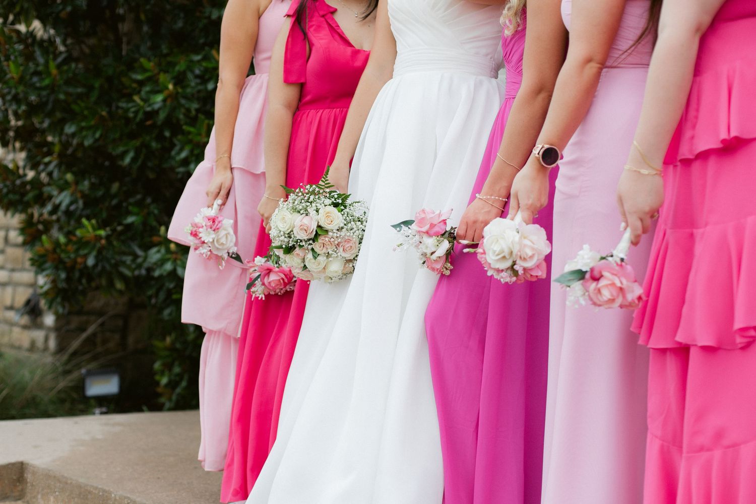 Bridesmaids in vibrant pink dresses stand together holding white bouquets at an outdoor wedding ceremony.