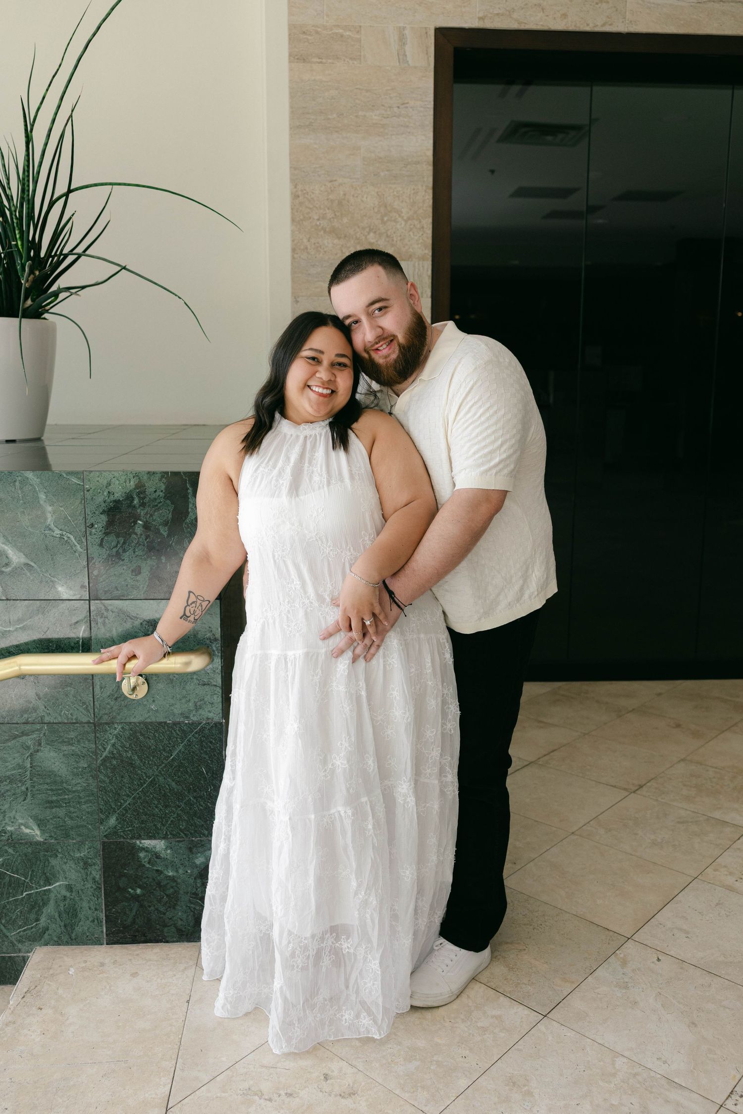 Couple in matching white outfits posing together near marble steps in an elegant indoor setting.