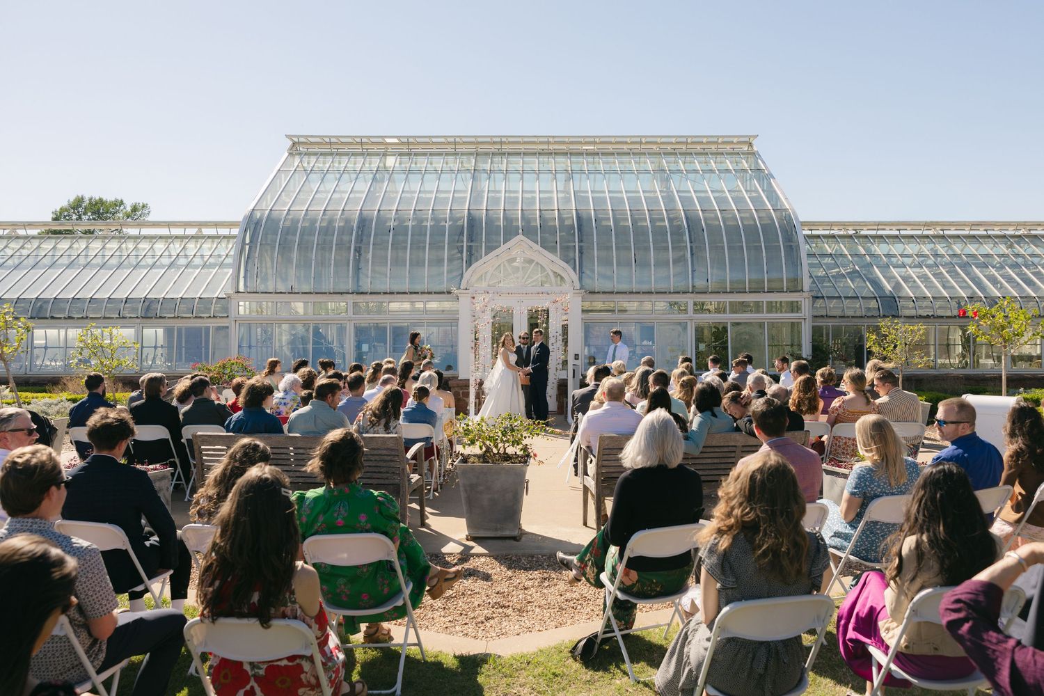 Outdoor wedding ceremony in front of a grand glass greenhouse conservatory with guests seated on white chairs.