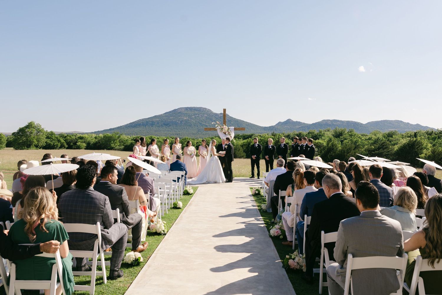 Outdoor wedding ceremony with guests seated in white chairs, aisle runner, and mountain backdrop under clear blue sky.