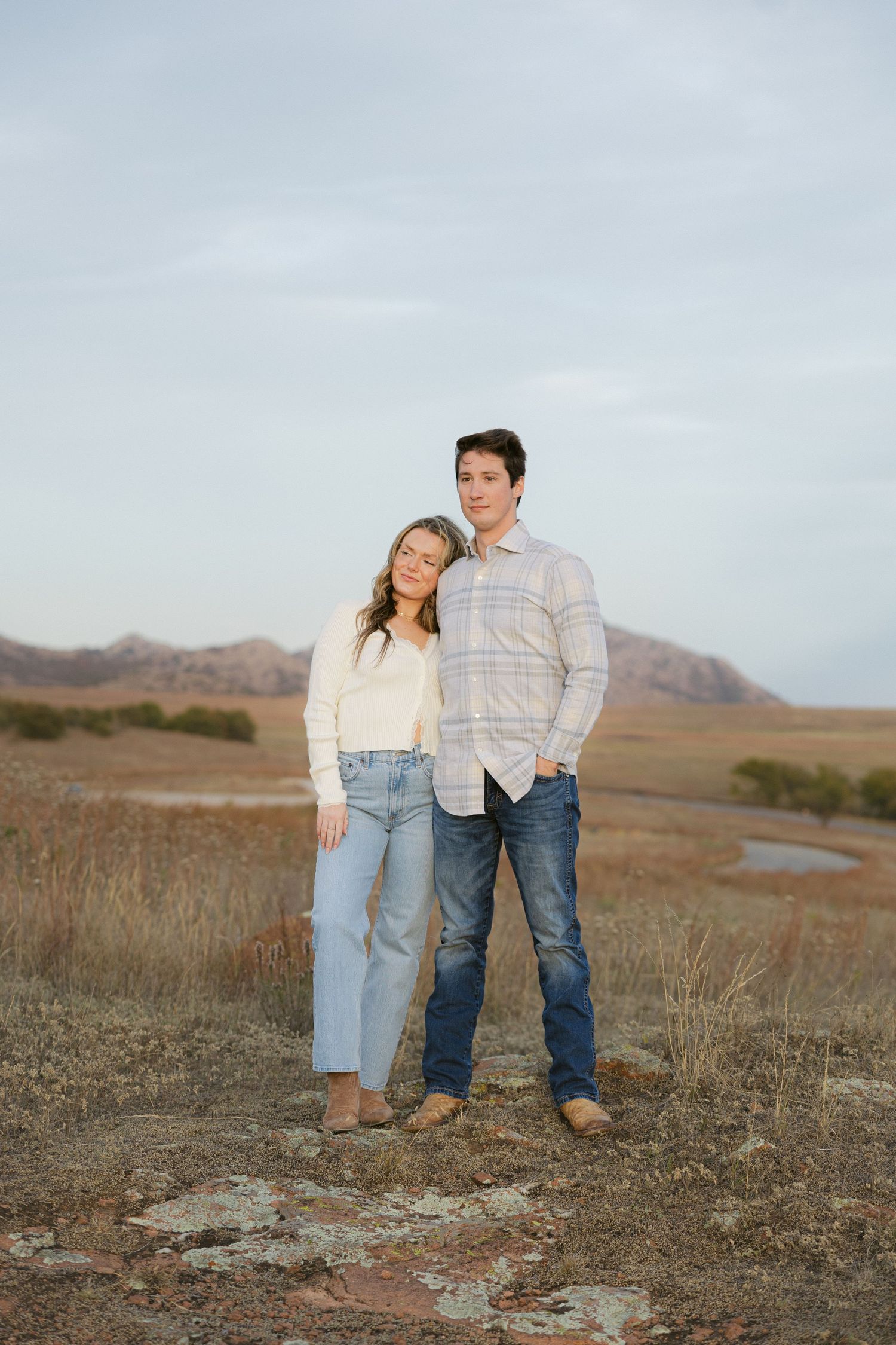 A couple stands together in an open field with mountains and rolling hills visible in the background during golden hour.
