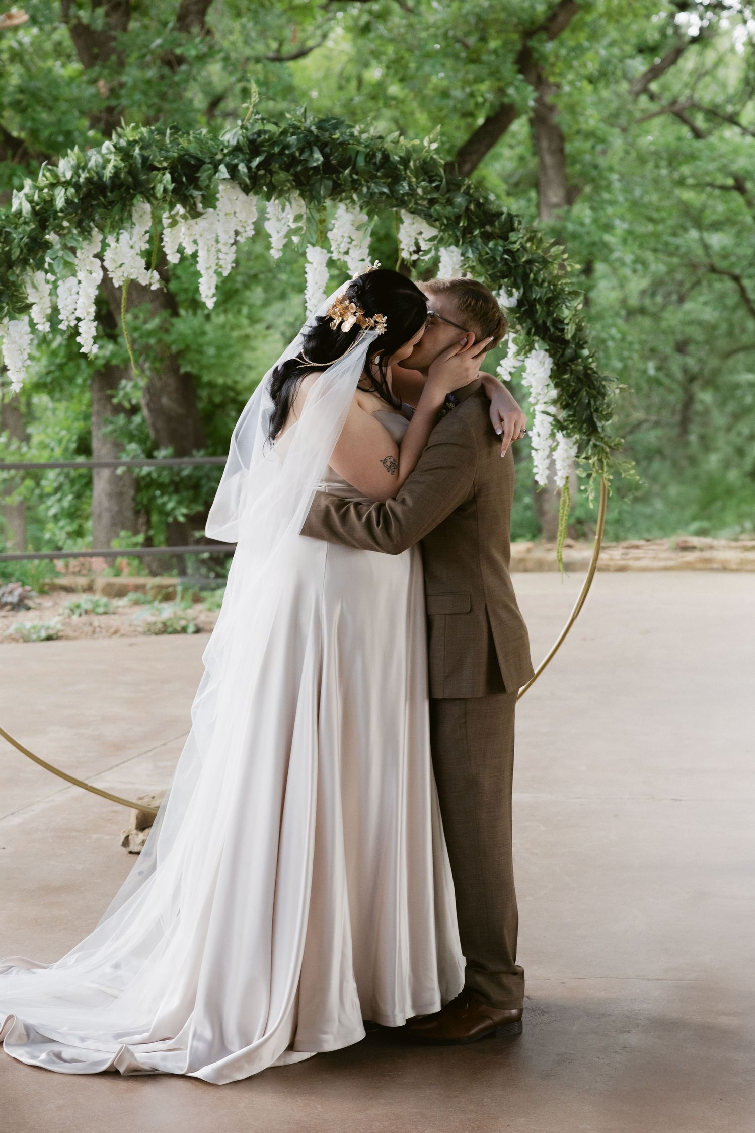 A romantic wedding moment under a floral arch with cascading white flowers in a garden setting.