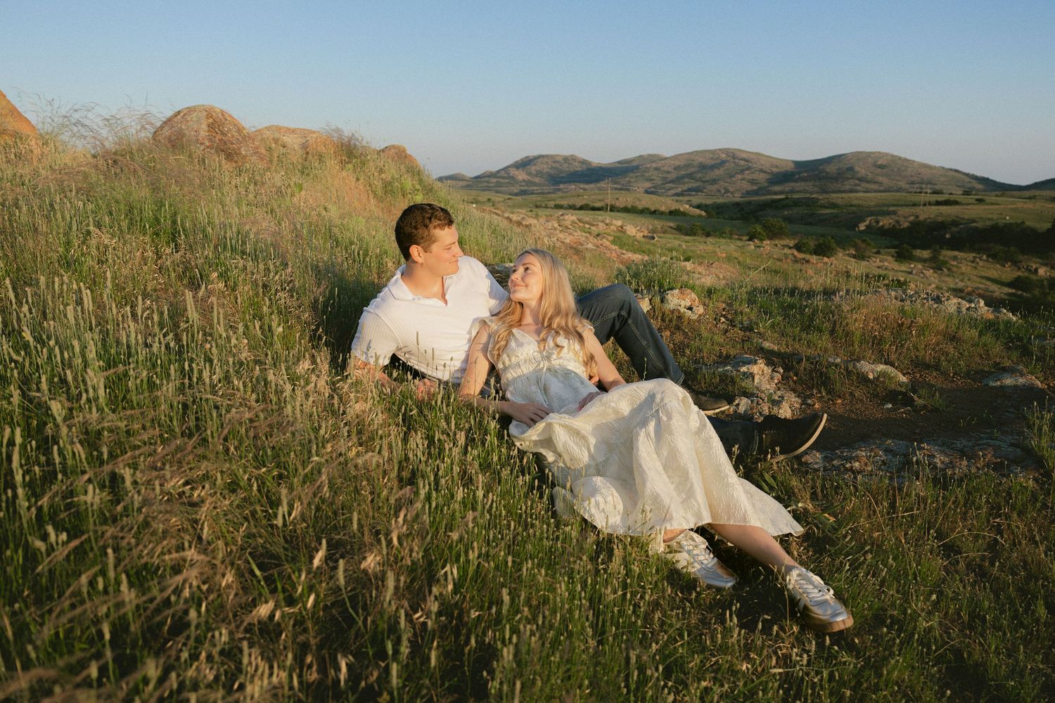 A couple embraces on a grassy hillside during golden hour with mountains in the background.