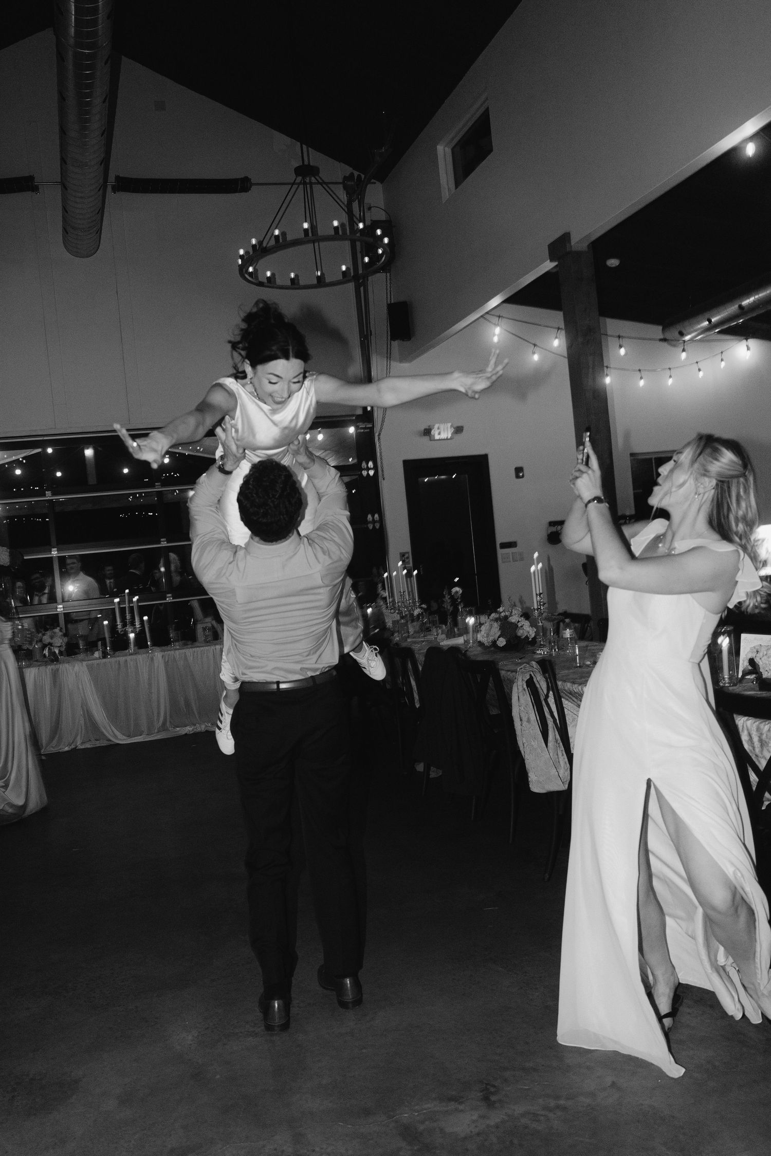 Black and white photo of groom lifting child while bride watches during fun wedding reception dance.
