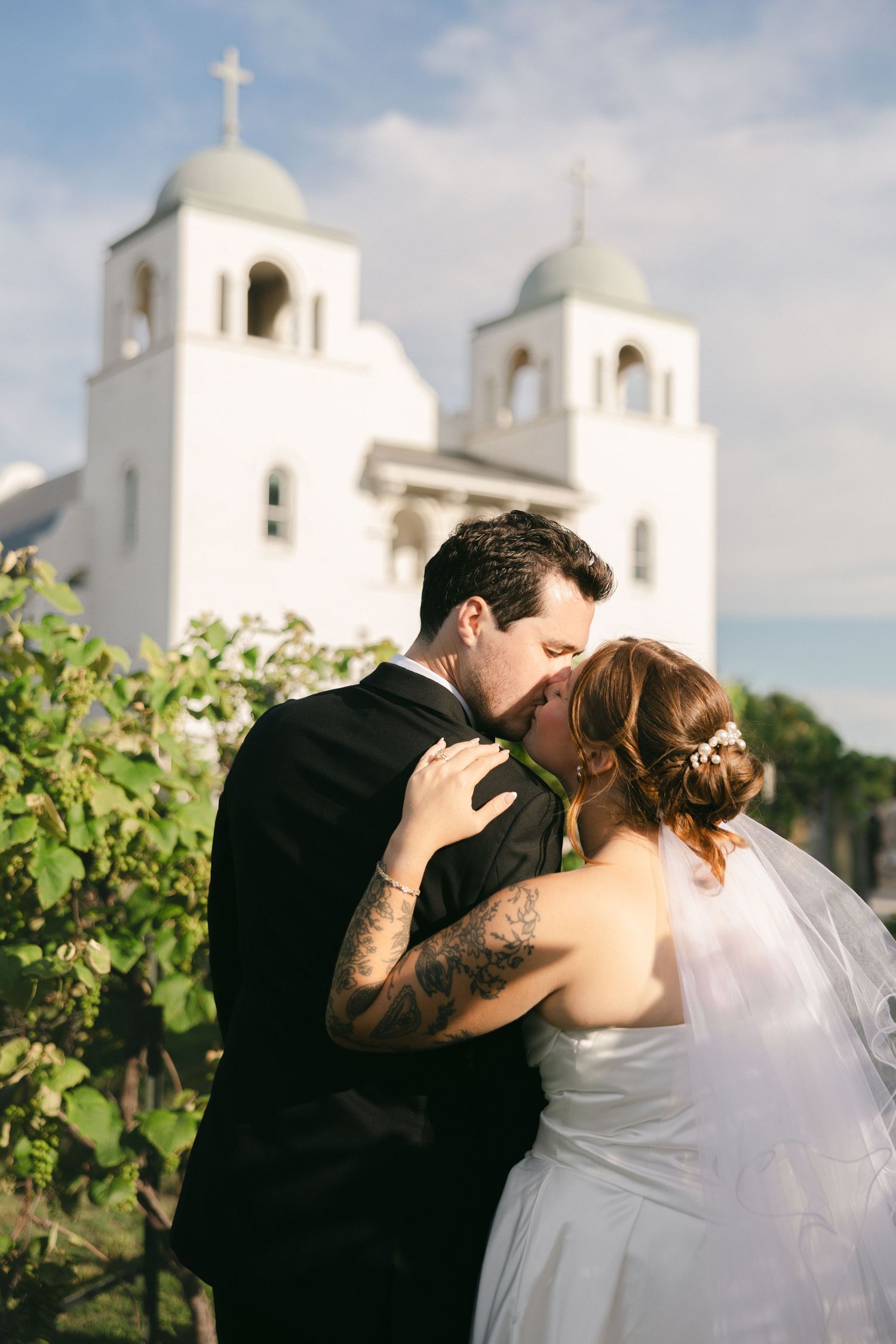 Bride and groom share a romantic kiss in front of a white church with green domes on their wedding day.