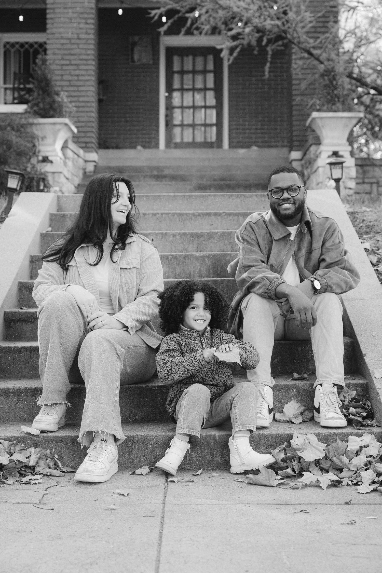Black and white photo of a happy family of three sitting on front porch steps outside a brick home.