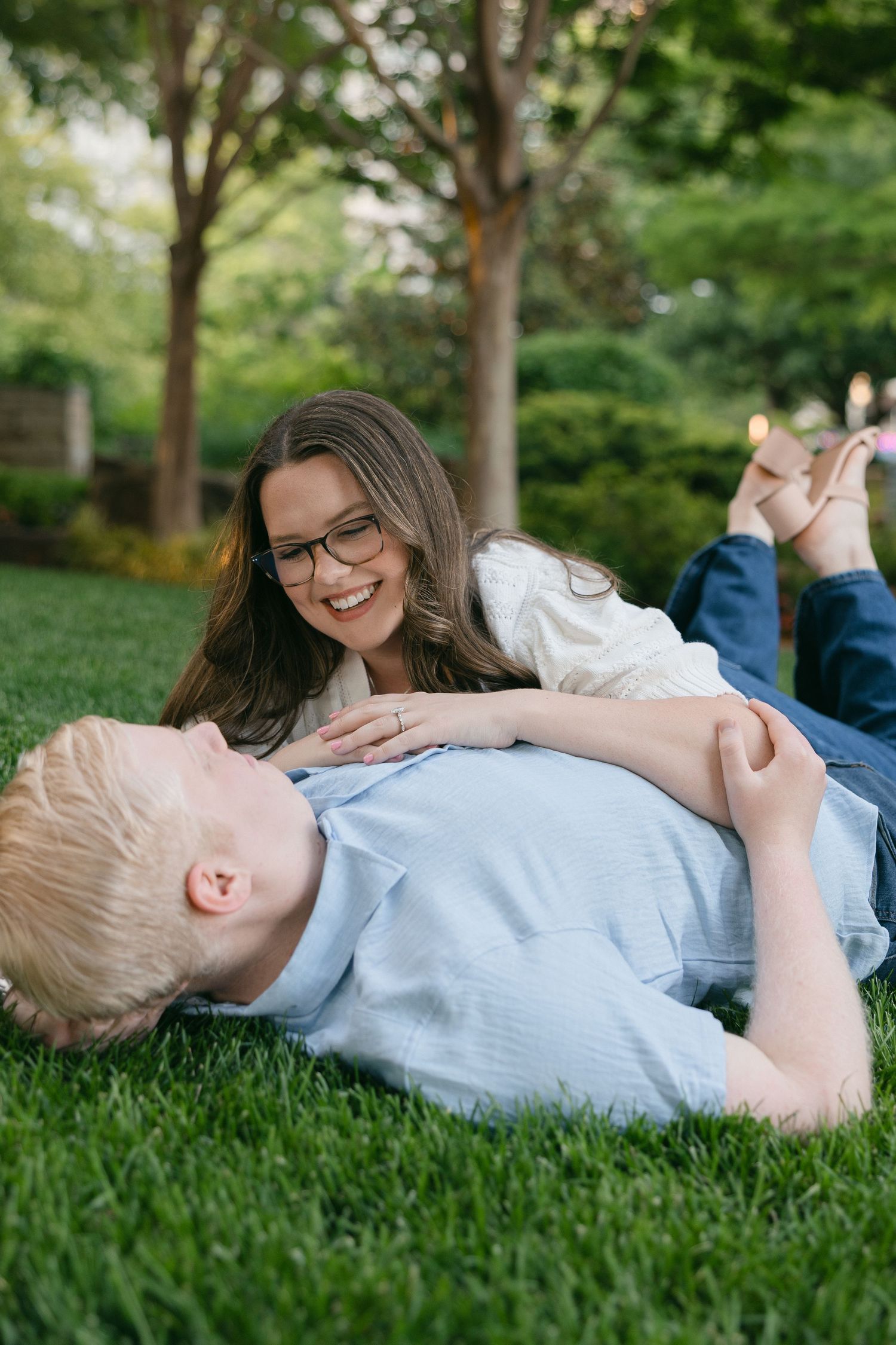 A smiling woman with glasses lies on green grass laughing with a blonde boy in a sunny park setting.