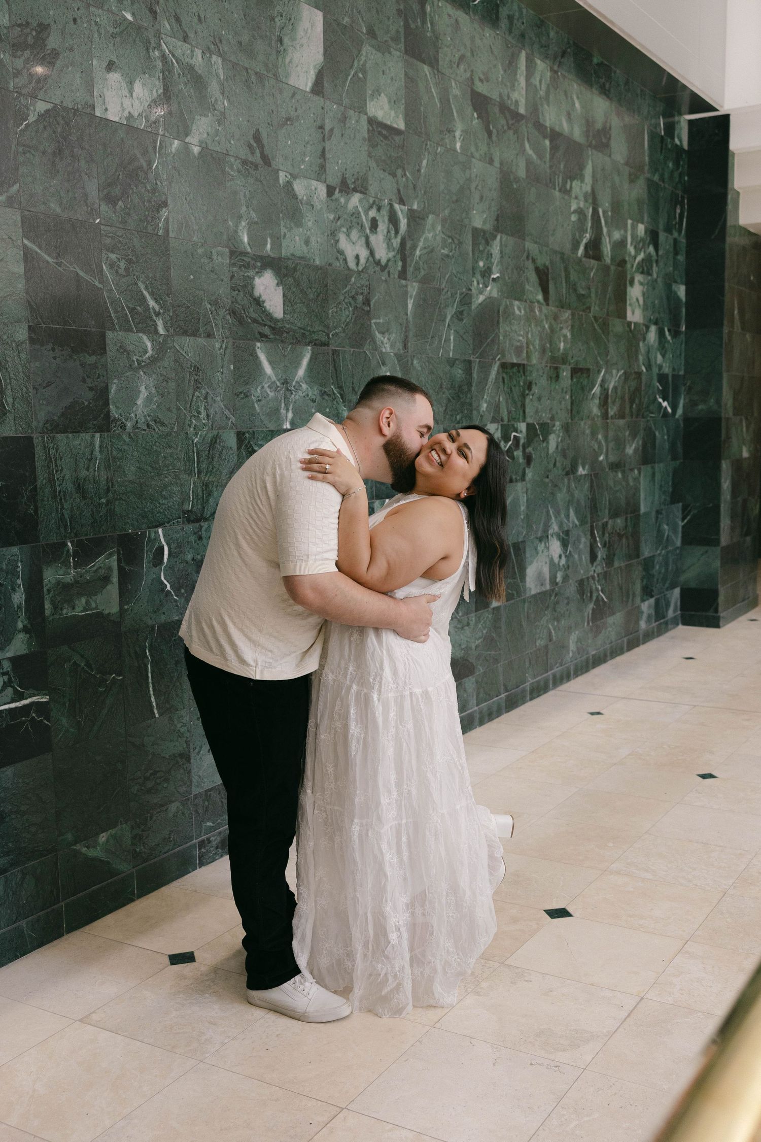Couple embracing and kissing in front of a green marble wall, woman in white dress smiling joyfully.