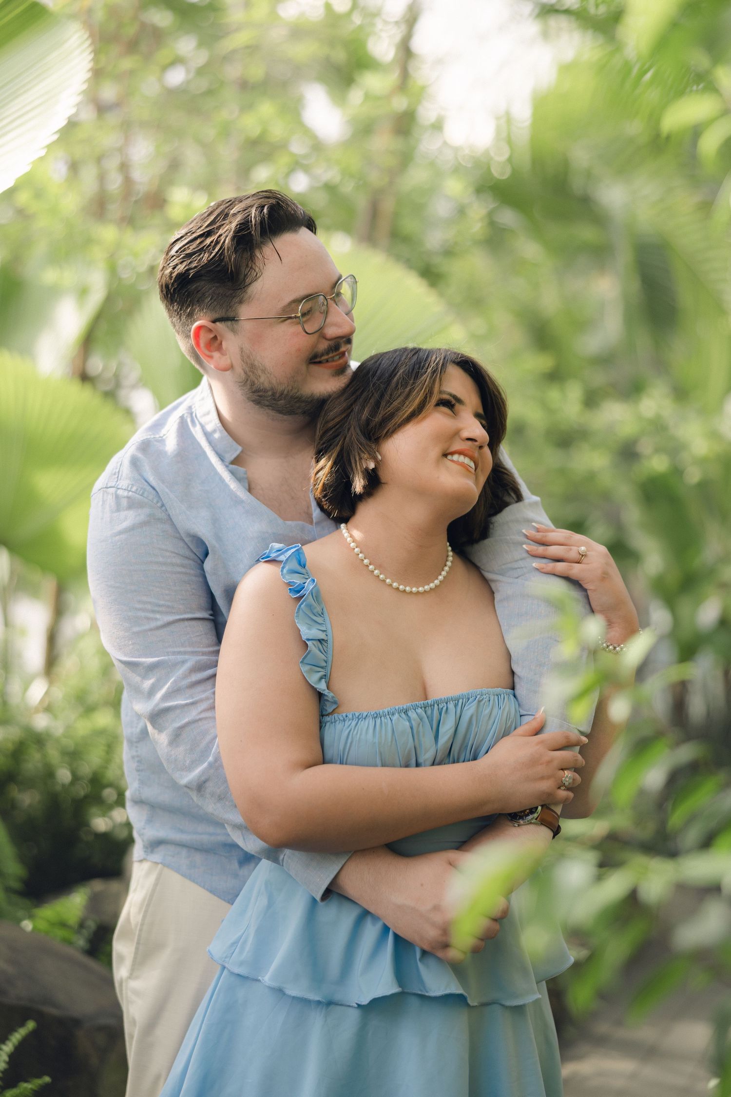Couple embracing in lush green garden, woman in blue dress with pearl necklace smiling, man in light blue shirt with glasses.