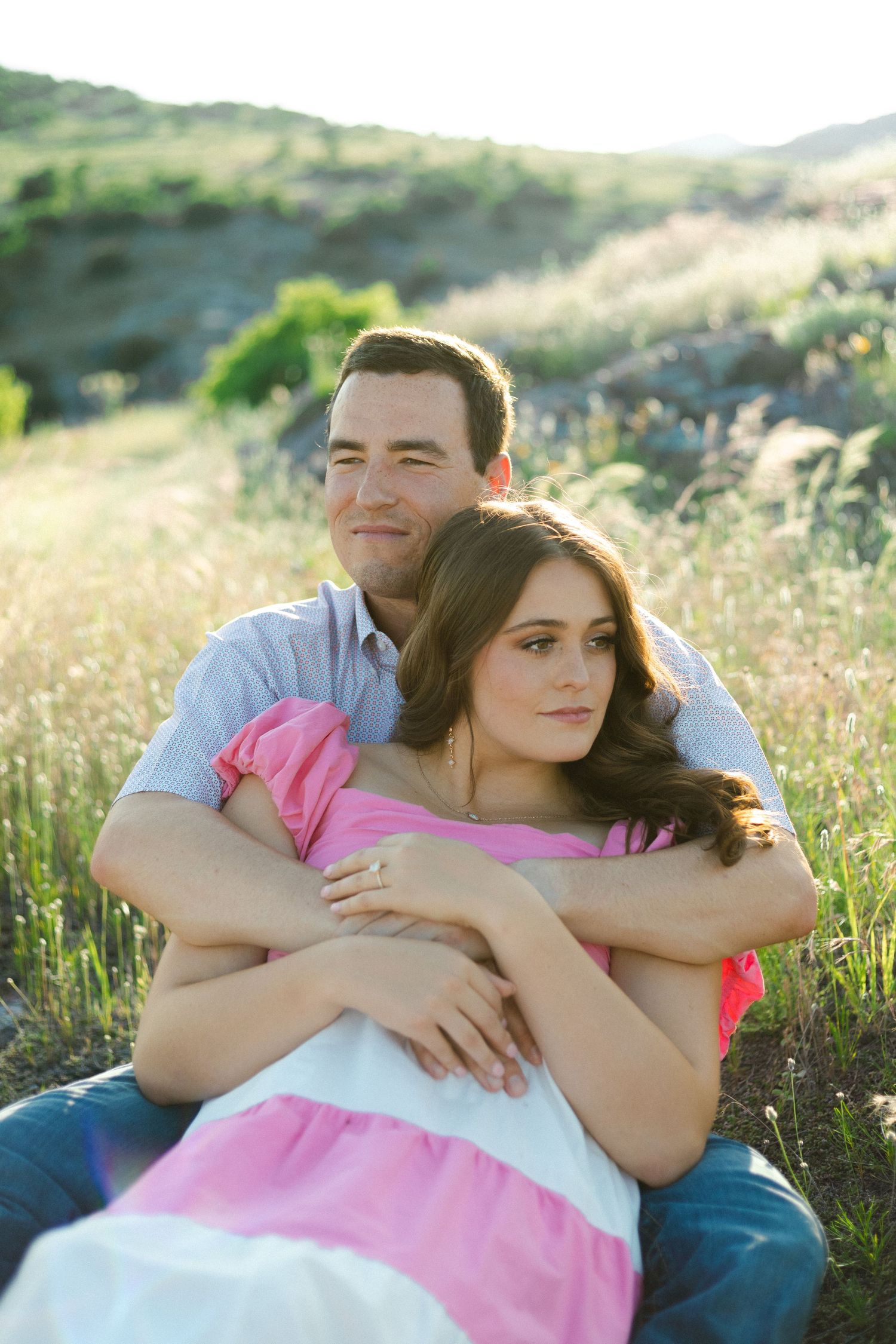 A couple sits together embracing in a grassy hillside meadow at golden hour with soft sunlight.
