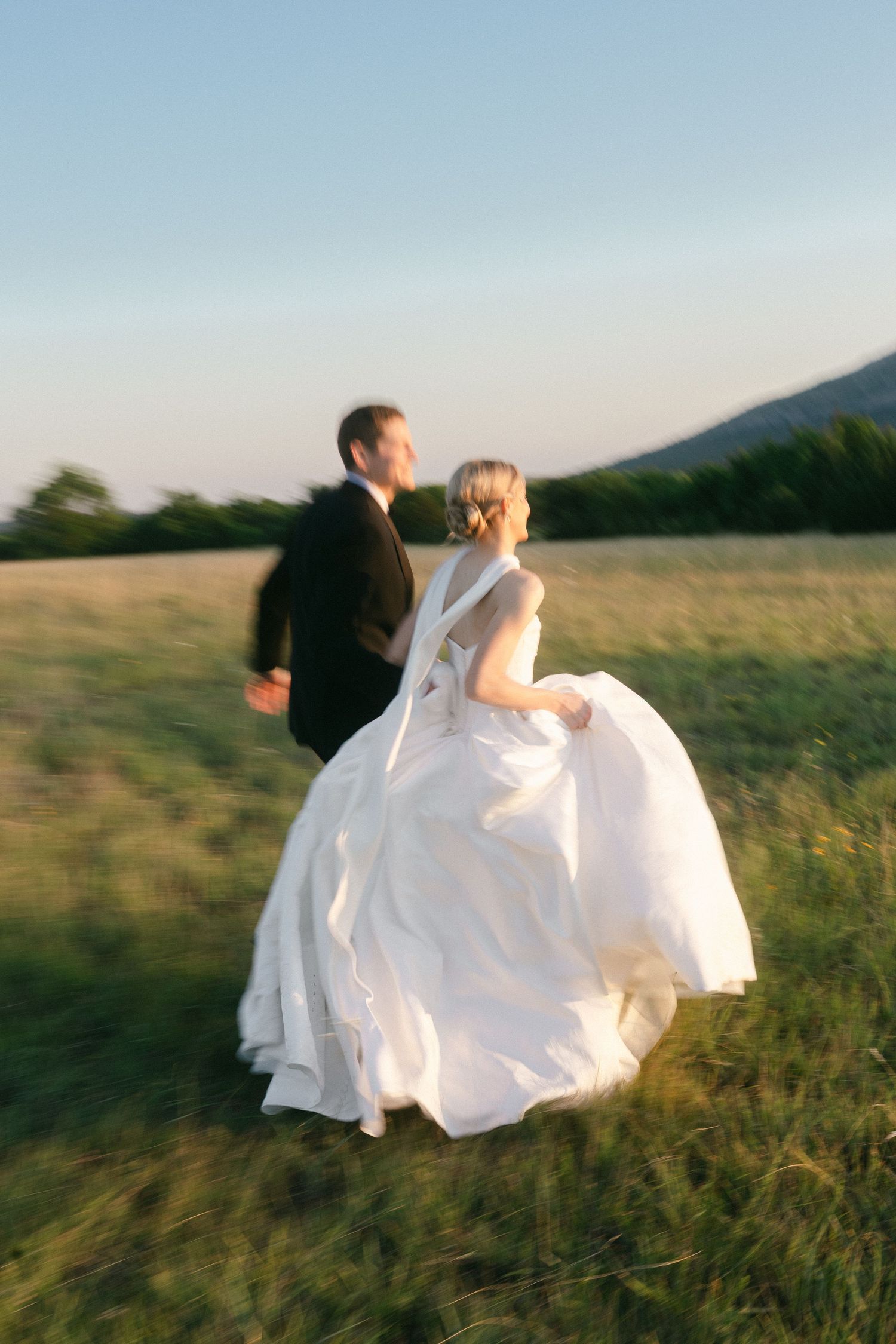 Bride in white ball gown and groom in black tux run through sunlit green field with mountain backdrop.