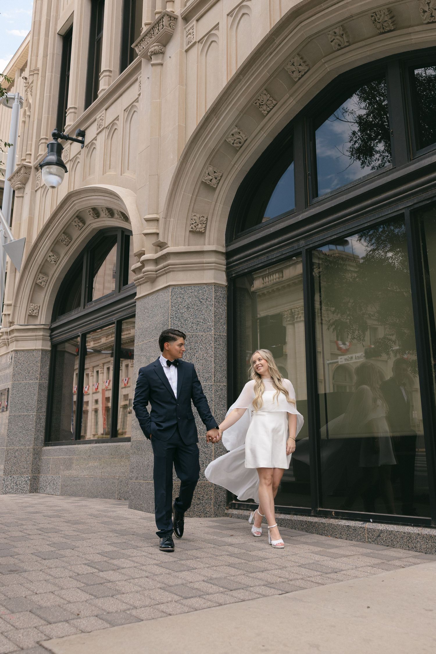 Couple in wedding attire holding hands walking past ornate beige stone building with arched windows on city sidewalk.