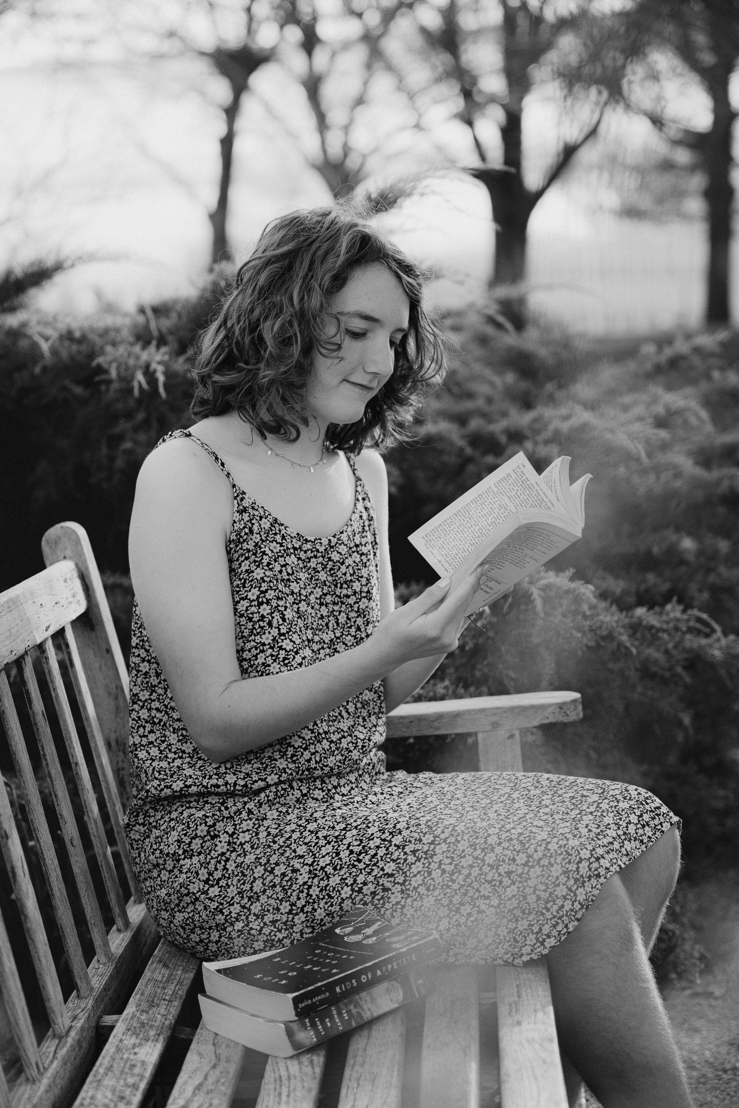 Black and white photo of someone reading a book while sitting on a wooden bench outdoors in a floral dress.