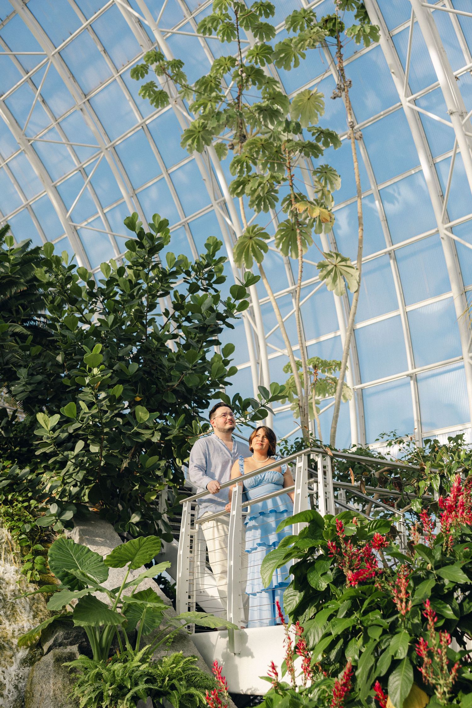 Couple standing on a white balcony platform inside a lush tropical greenhouse with palm trees and a glass dome ceiling.