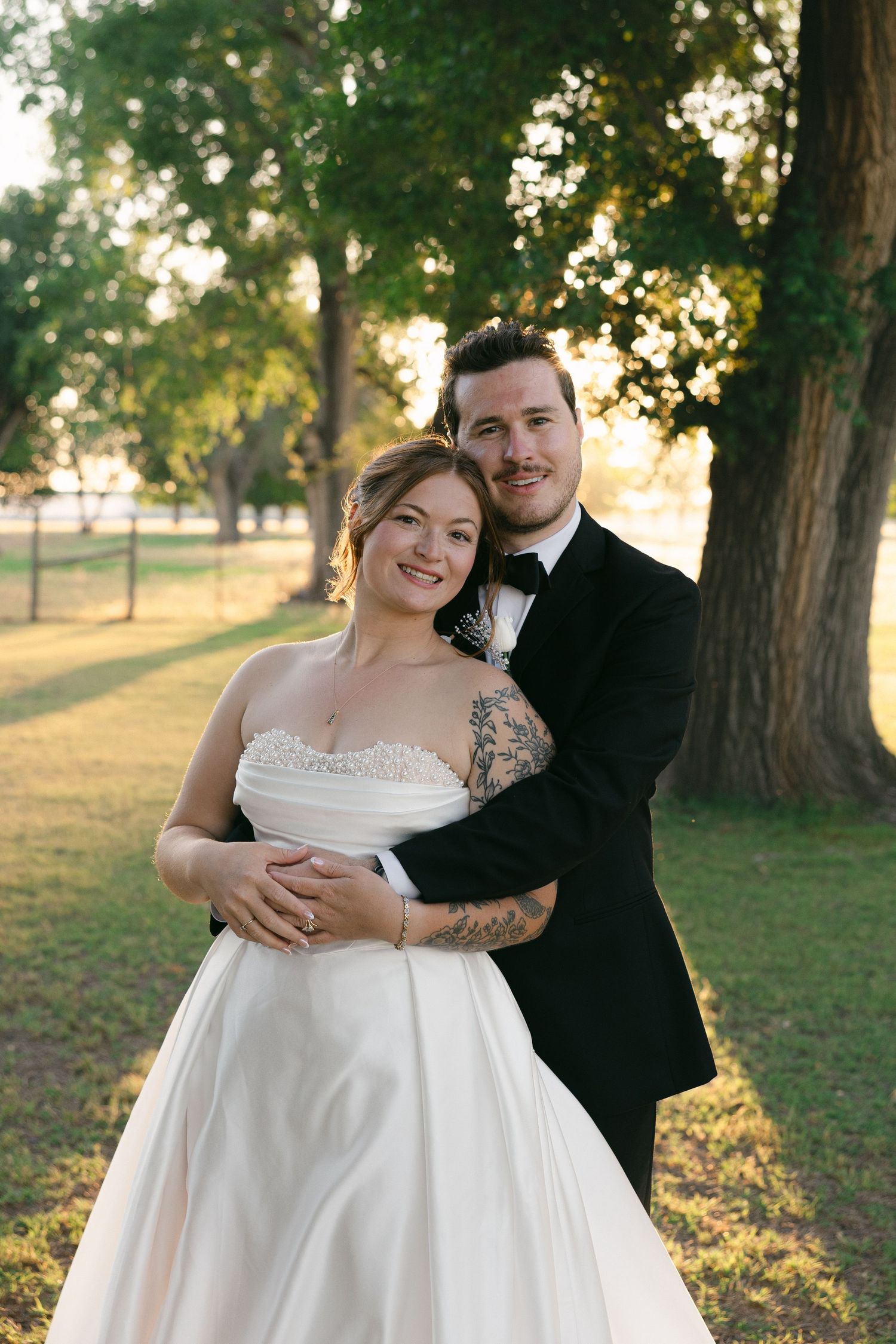 Bride in strapless white gown and tattooed groom in black tuxedo embrace outdoors at golden hour.