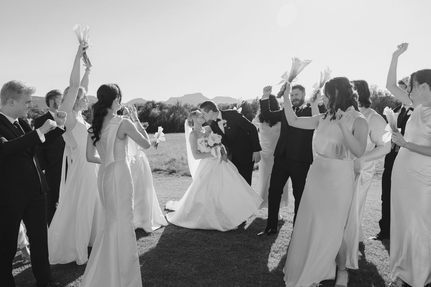 Black and white wedding photo of bride and groom kissing surrounded by cheering bridal party outdoors.