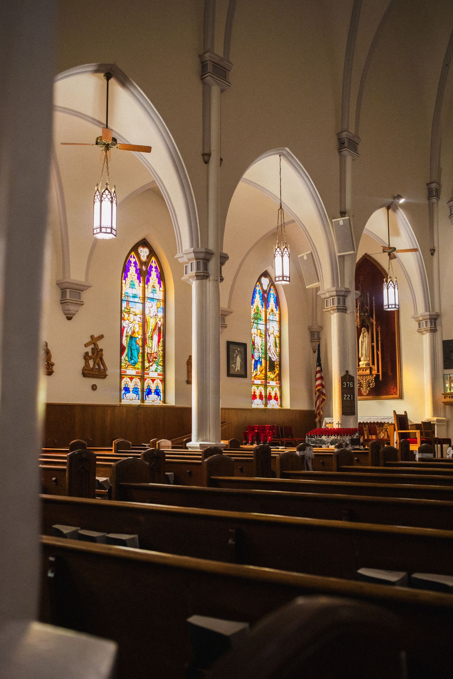 Interior of a Catholic church with stained glass windows, wooden pews, and Gothic arches illuminated by pendant lighting.