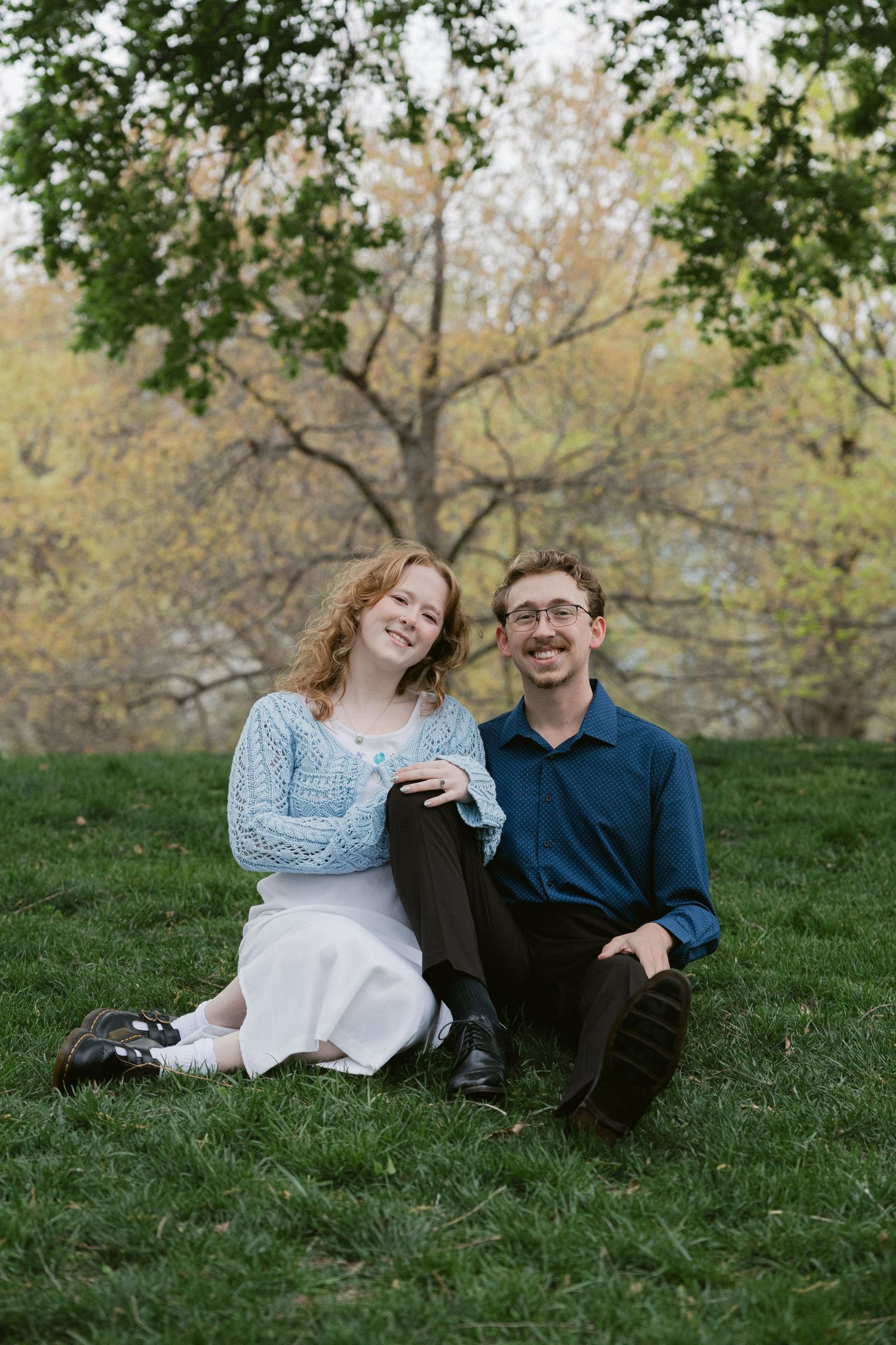 Couple sitting together on grass with trees in background during outdoor photo session.