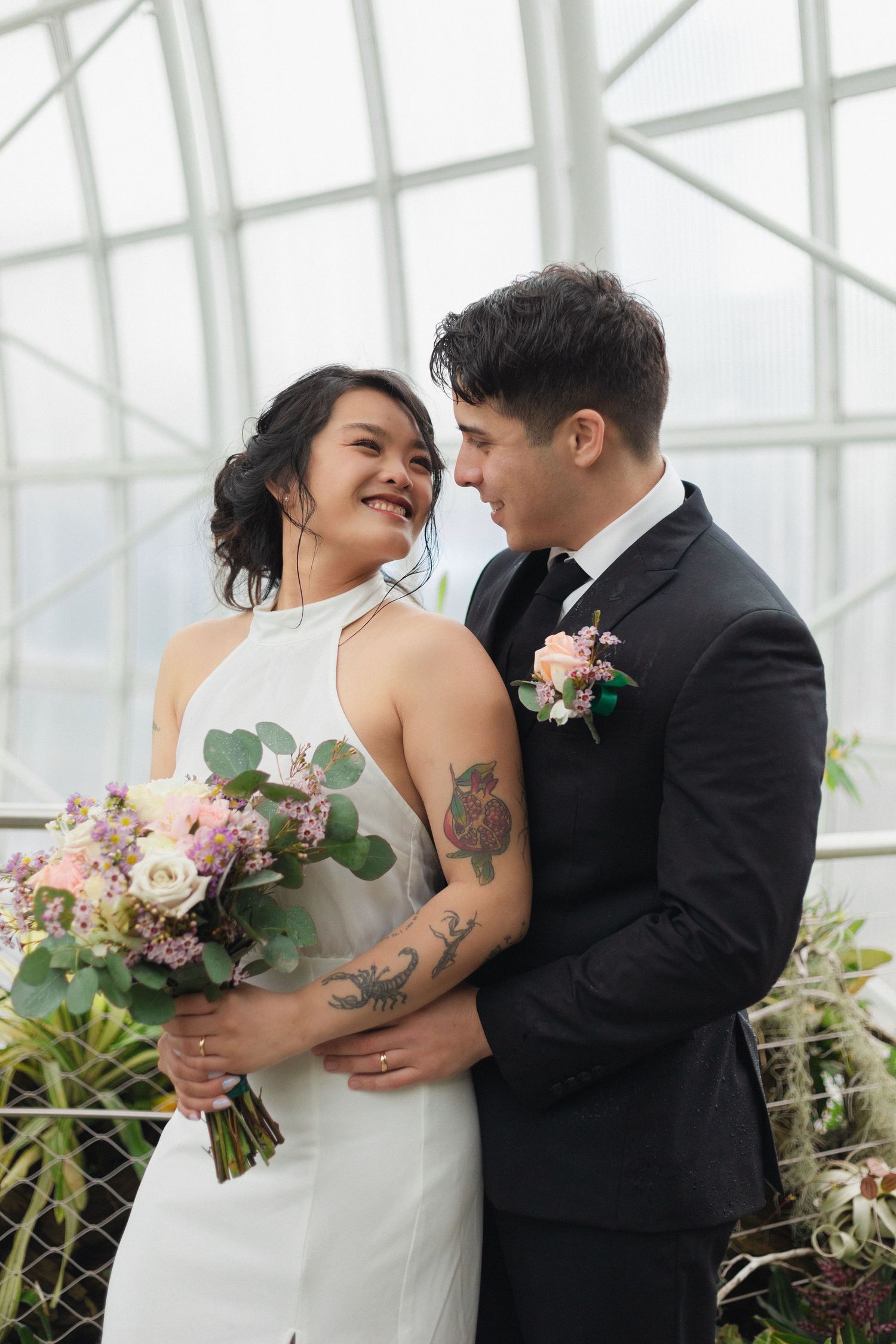 A romantic wedding portrait of newlyweds in a greenhouse, with bride holding a pink floral bouquet.