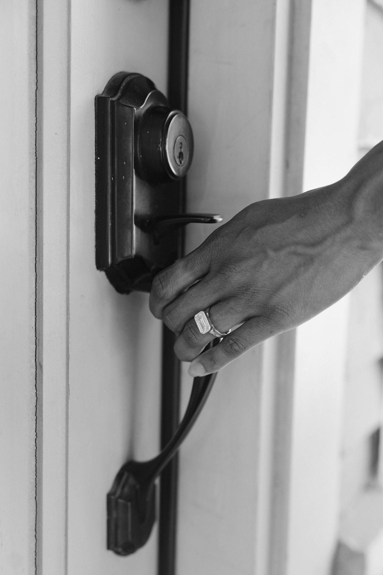 Black and white photo of a hand wearing a ring turning a door handle lock.