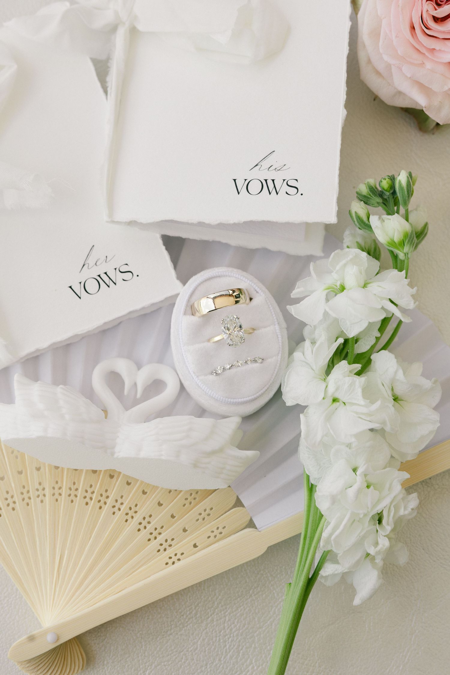 Elegant wedding flat lay with vow books, gold rings in velvet box, swan figurines, white fan, and fresh flowers.
