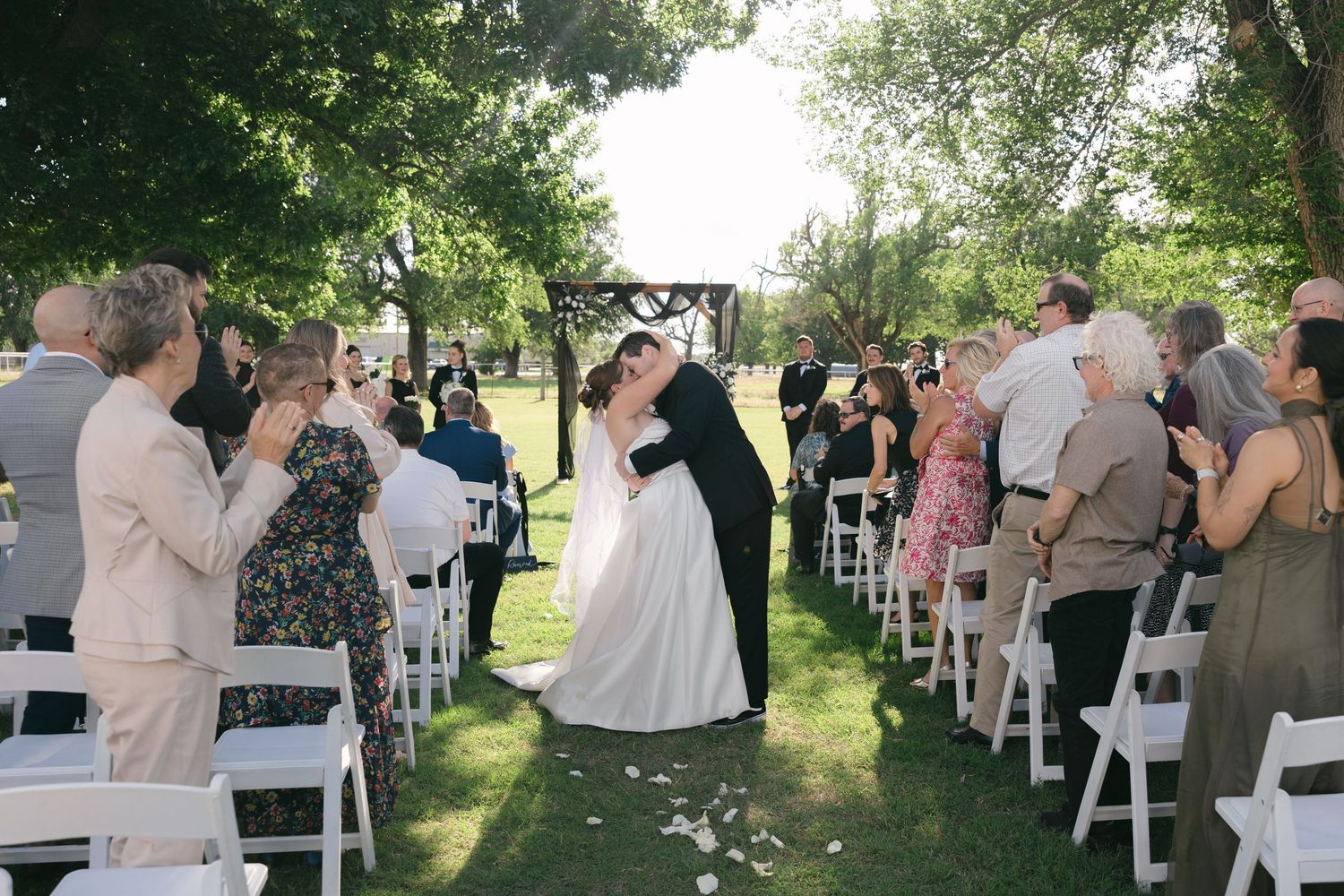 Bride and groom share a kiss at outdoor wedding ceremony with guests applauding along white chair-lined aisle under green trees.