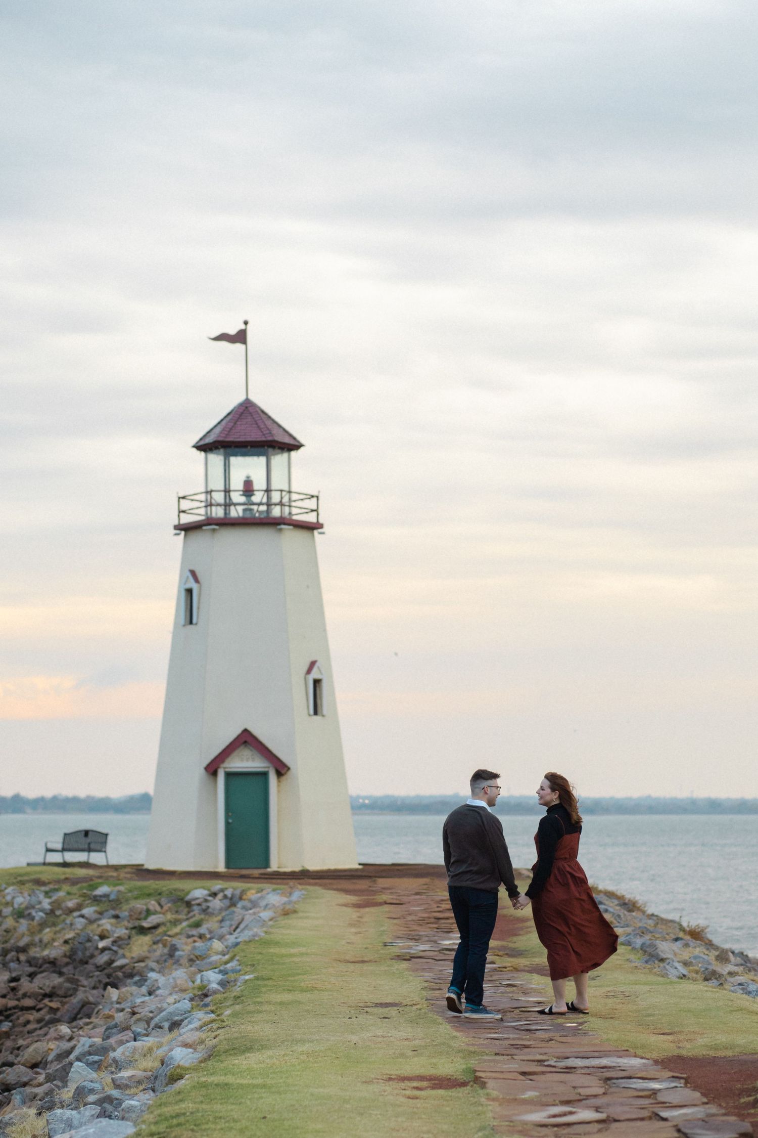 A couple stands on a rocky path near a white lighthouse with a green door at dusk.