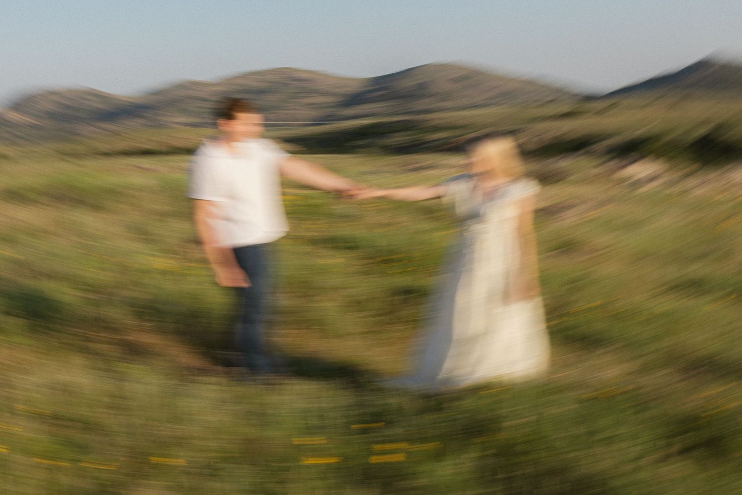 Two silhouettes walk hand in hand through rolling green hills at sunset creating a romantic countryside scene.