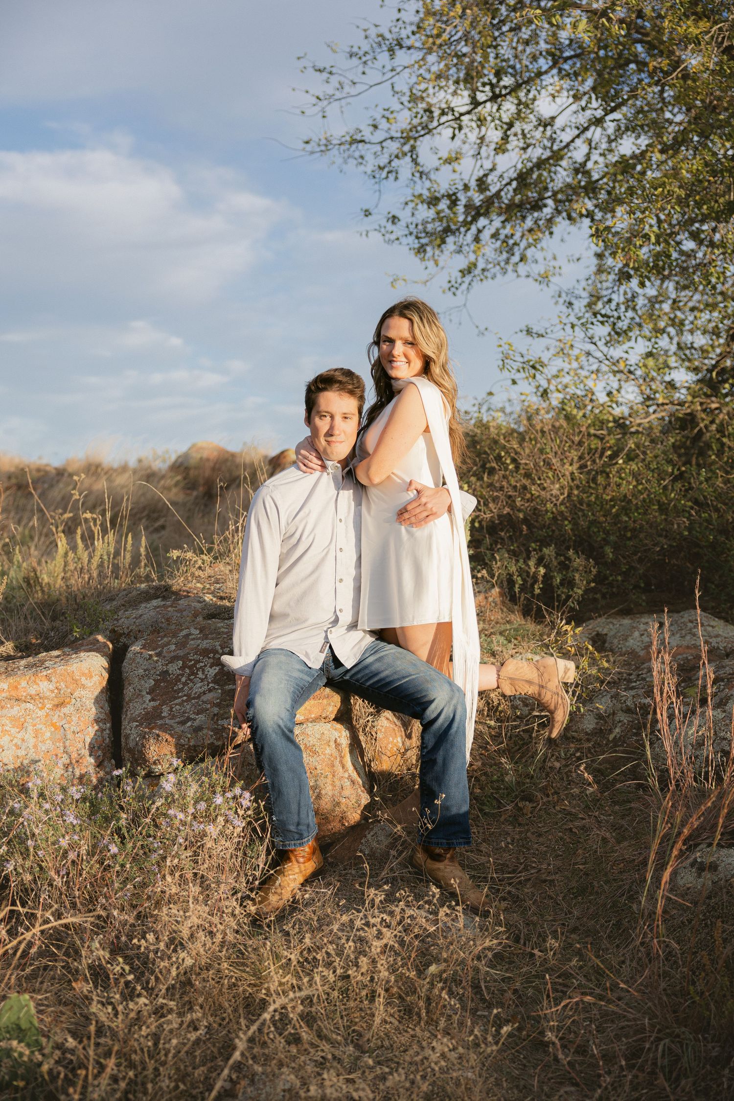 Couple sitting together on rocky terrain during golden hour with desert brush and trees in background.